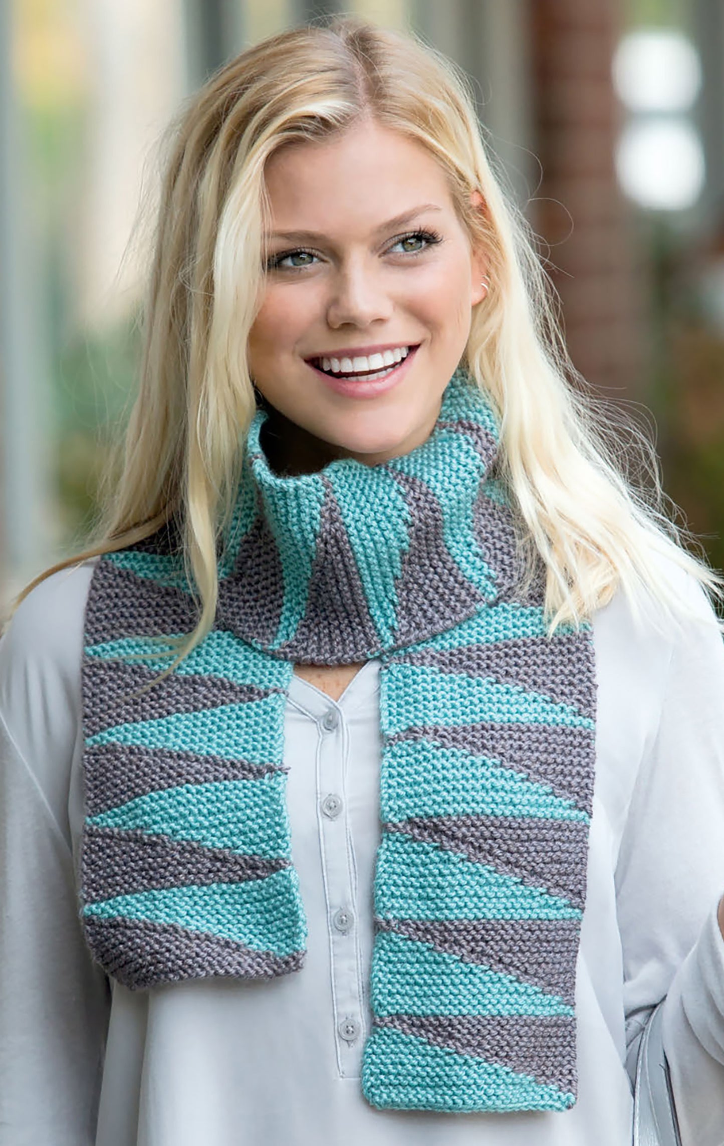 A smiling woman with long blonde hair wears a light gray shirt and a turquoise-and-gray striped scarf from the "Warm up with Scarves & Cowls" collection by Leisure Arts, standing in a softly blurred indoor setting.