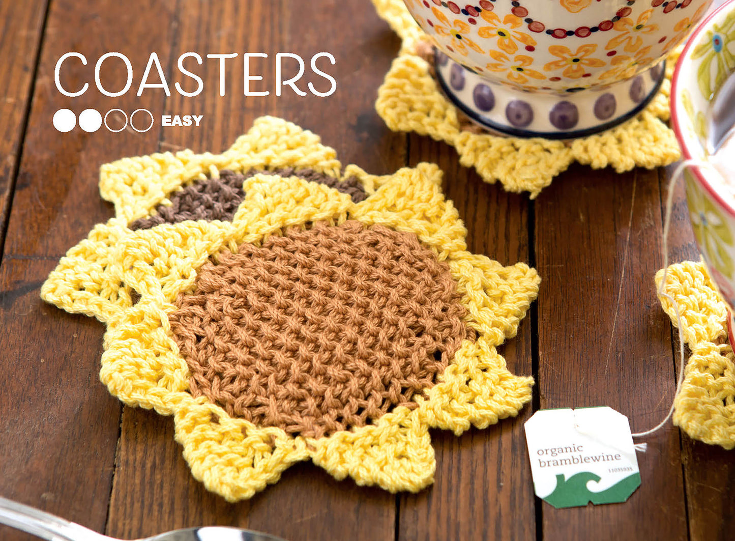 A Leisure Arts book, Stitch A Garden—18 Designs Charted for Beading and Cross Stitch, rests on a wooden table near colorful thread and embroidery hoops. Ideal for cross stitch lovers. Text reads STITCHING with an easy difficulty rating.