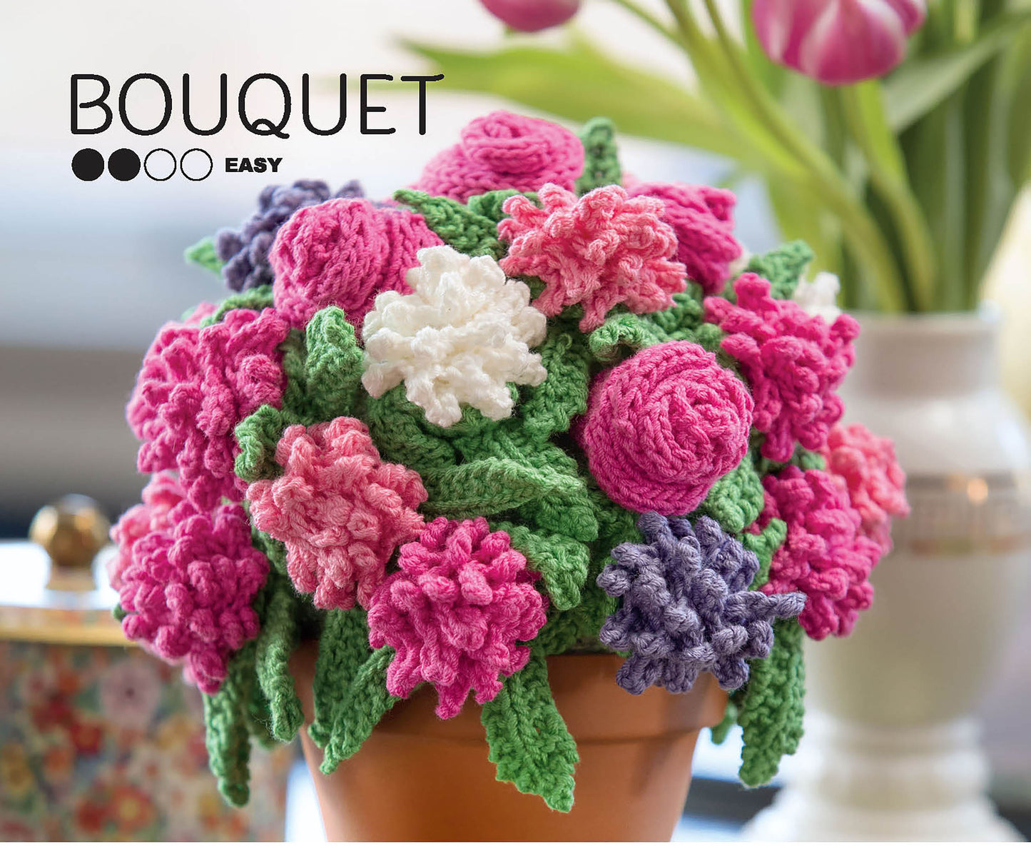 A copy of "Stitch A Garden—18 Designs Charted for Beading and Cross Stitch" by Leisure Arts is shown. The cover features bright crocheted flower bouquets in a terracotta pot, with four black circles and blurred tulips in the background.