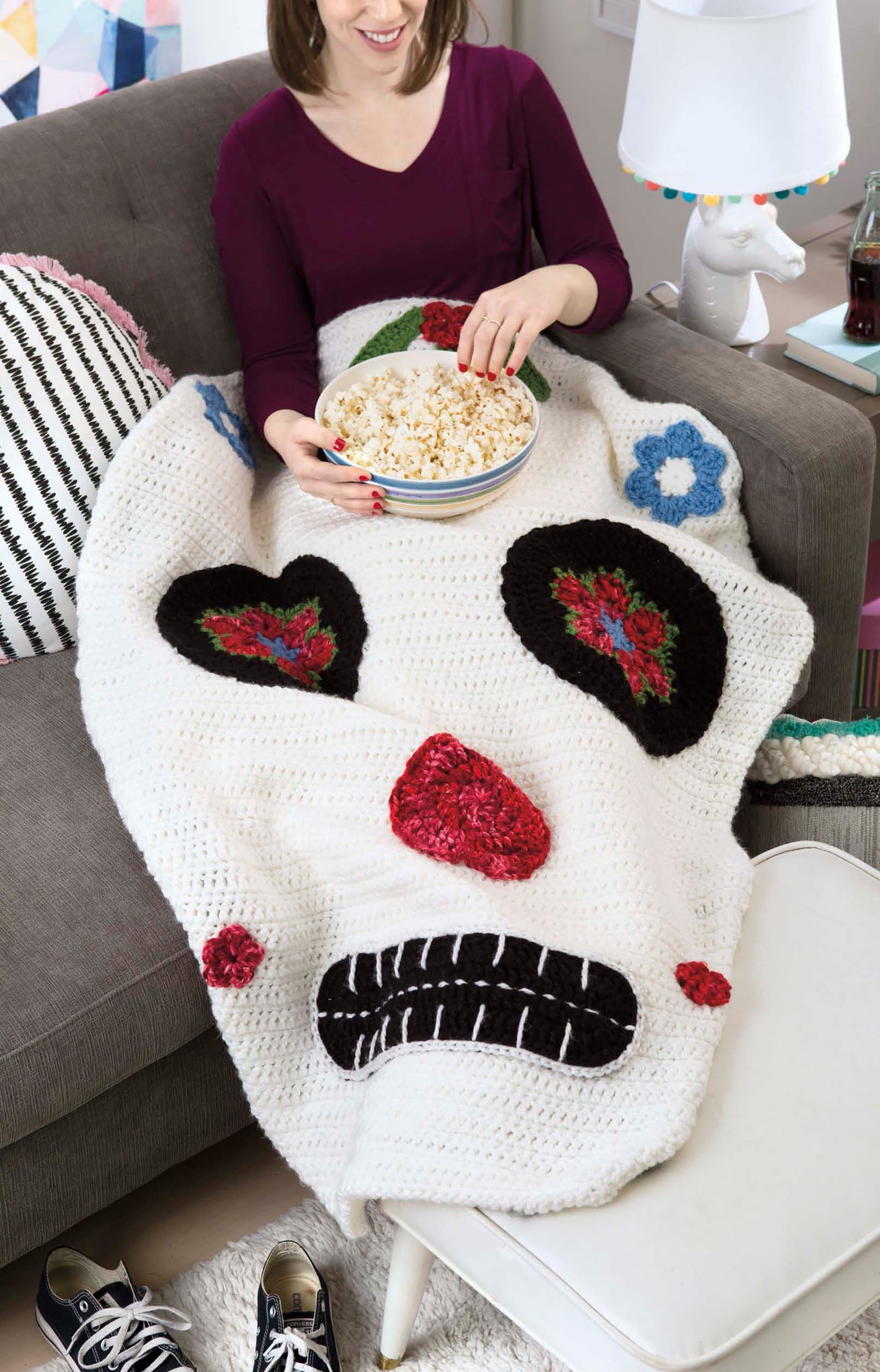 A woman smiles on a couch with her legs wrapped in a Leisure Arts Kids' Snuggies shaped like a sugar skull. She holds popcorn, and a soda bottle plus shoes are nearby.