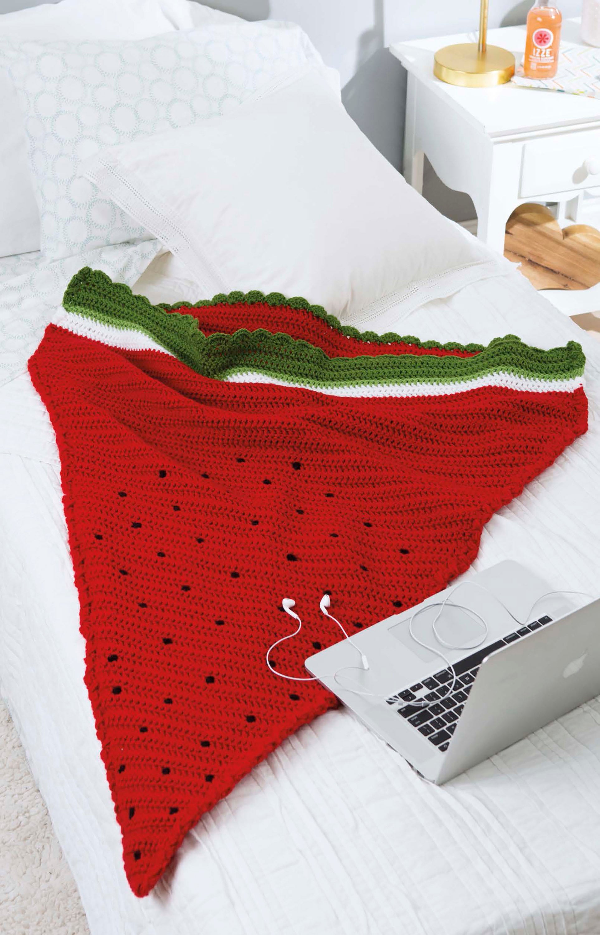 The Leisure Arts Kids' Snuggies blanket, designed like a watermelon slice in red, white, and green, rests on a bed beside a laptop and earphones—ideal for fans of cozy kids' blankets with fun shapes.