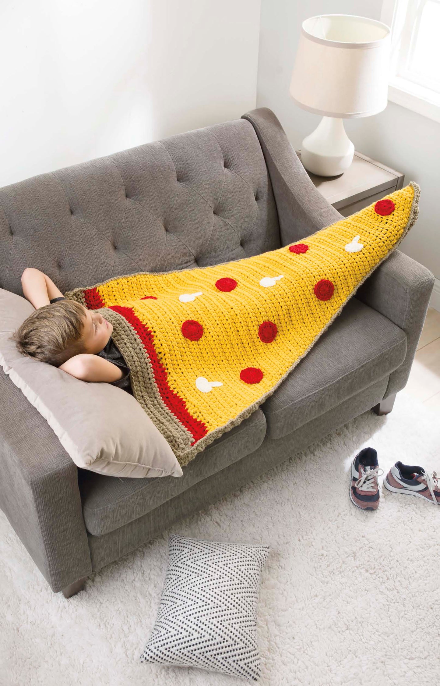 A child lounges on a gray sofa, wrapped in a Leisure Arts Kids' Snuggies shaped like a giant pizza slice with red and white toppings. Shoes rest on the floor close by, and a lamp is placed on a table near the window.
