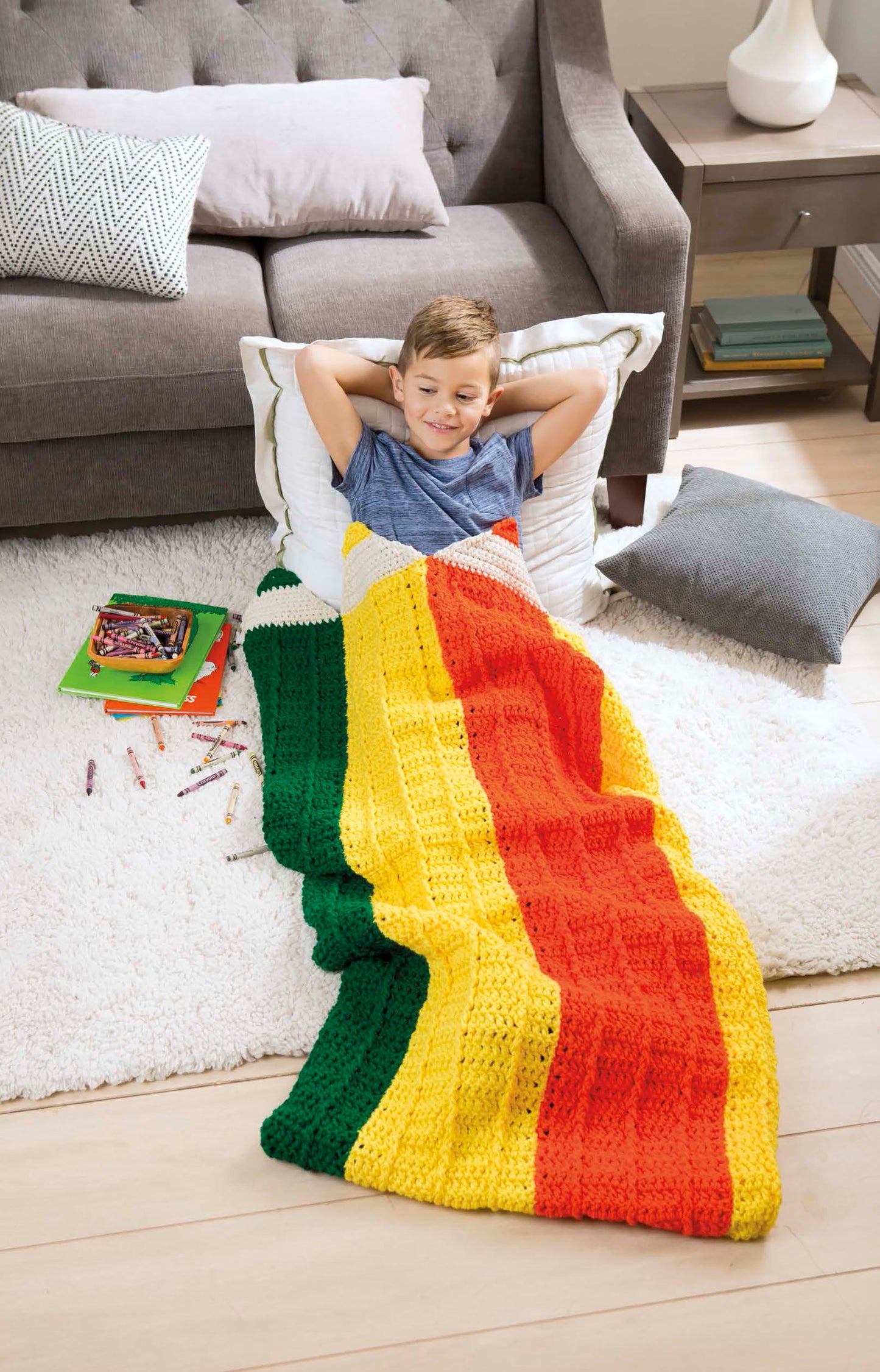 A smiling child lies on a white rug, wrapped in a Leisure Arts Kids' Snuggies blanket shaped like a pencil. Crayons and paper are scattered nearby, with a gray sofa and table in the background.
