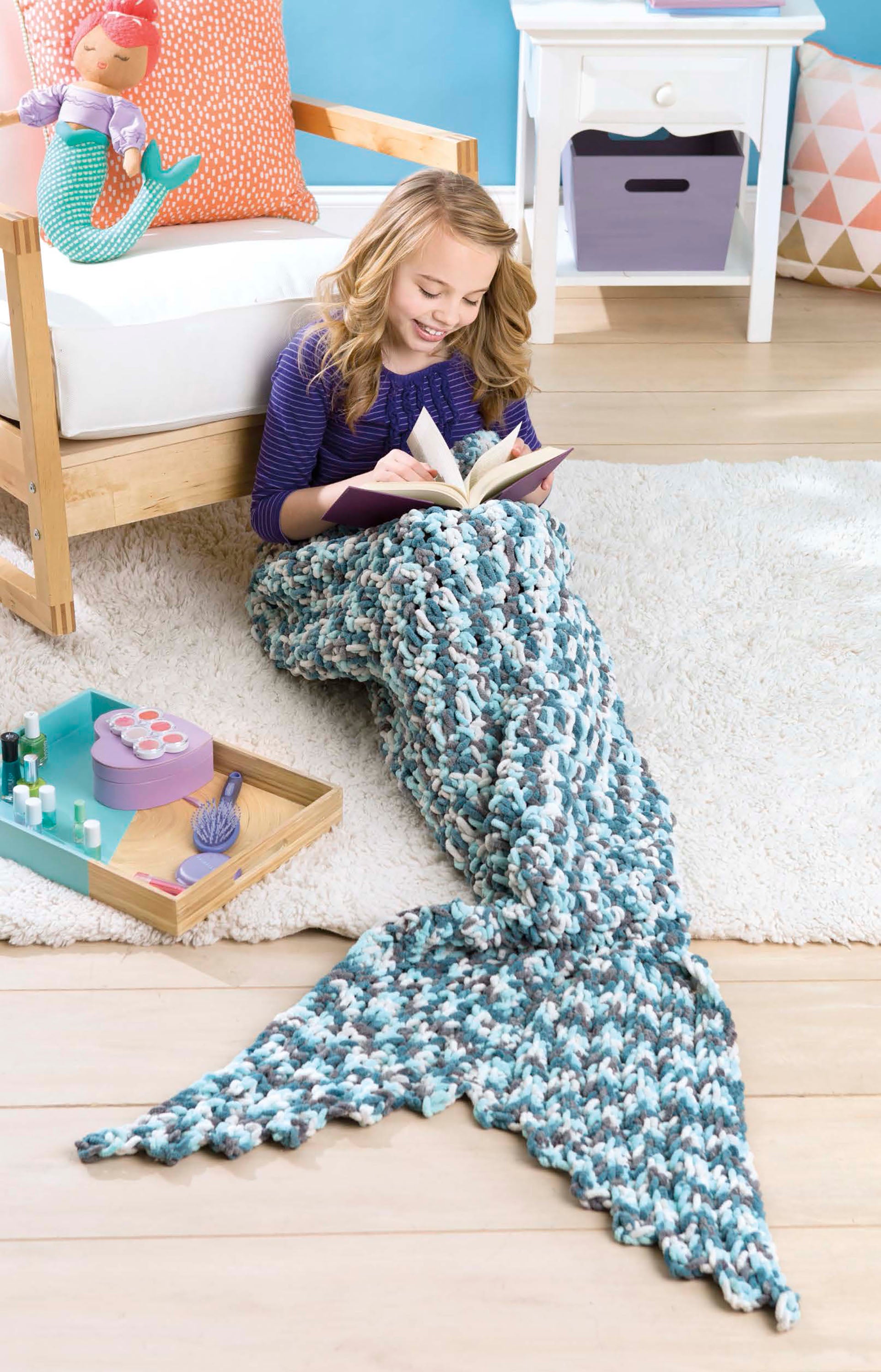 A girl sits on the floor reading, wrapped in a blue and white Leisure Arts Kids' Snuggies mermaid tail blanket. Nearby are a tray with nail polish and yarn, and a mermaid doll pillow rests on the couch behind her.