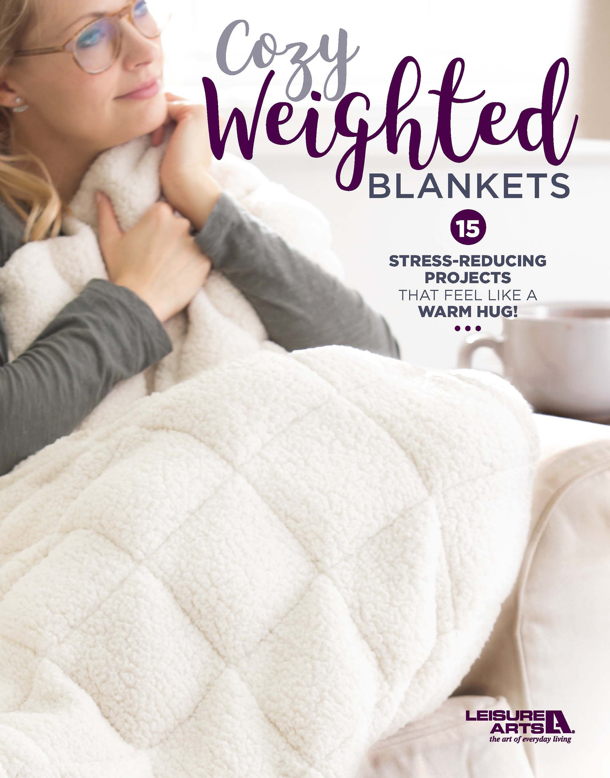 A woman with glasses sits on a couch, holding a fluffy white weighted blanket. Cozy Weighted Blankets by Leisure Arts offers 15 calming, stress-reducing sleep projects for soothing comfort—like a warm hug!.