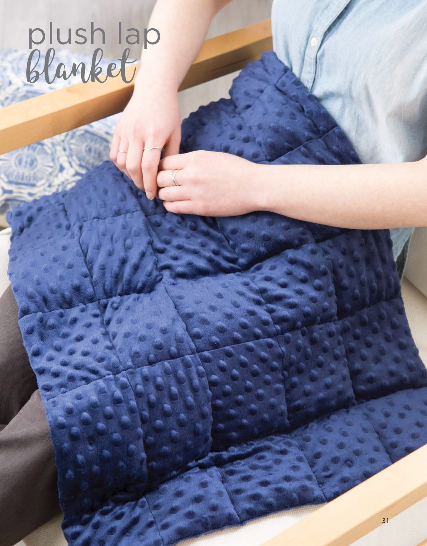 A person sits in a light chair with a dark blue, textured Cozy Weighted Blanket from Leisure Arts draped over their legs. Their hands rest on the blanket. The text "plush lap blanket" appears in the top left corner.