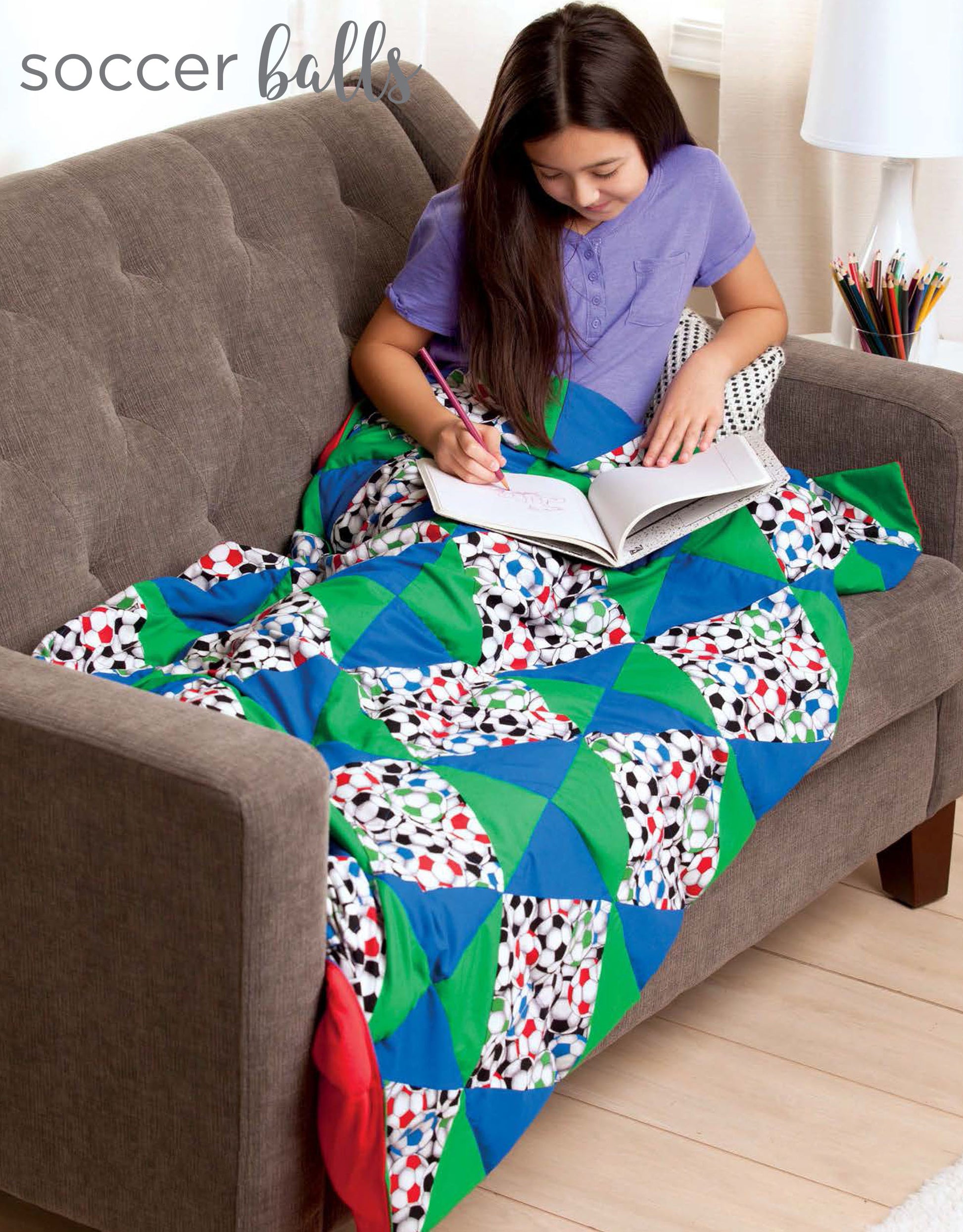 A girl relaxes on a gray couch, wrapped in a Leisure Arts Cozy Weighted Blanket with soccer ball patterns, drawing in her notebook. Colored pencils sit in a holder on the nearby table.