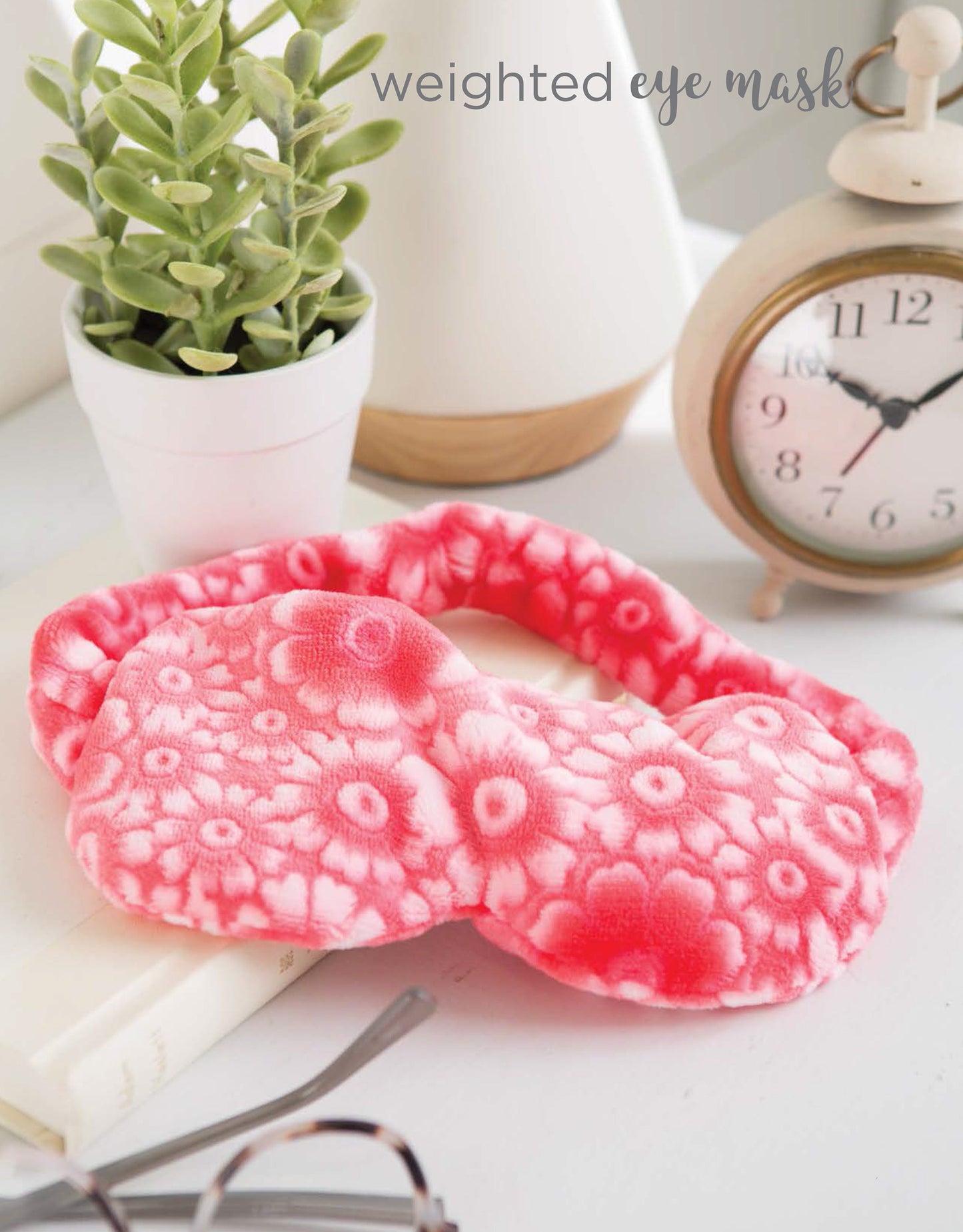 A soft, pink floral-patterned Cozy Weighted Blanket by Leisure Arts sits on a white book beside a potted plant, beige lamp, and vintage alarm clock at 7:09—perfect for calming relaxation and sleep. "Cozy Weighted Blanket" is displayed at the top right.