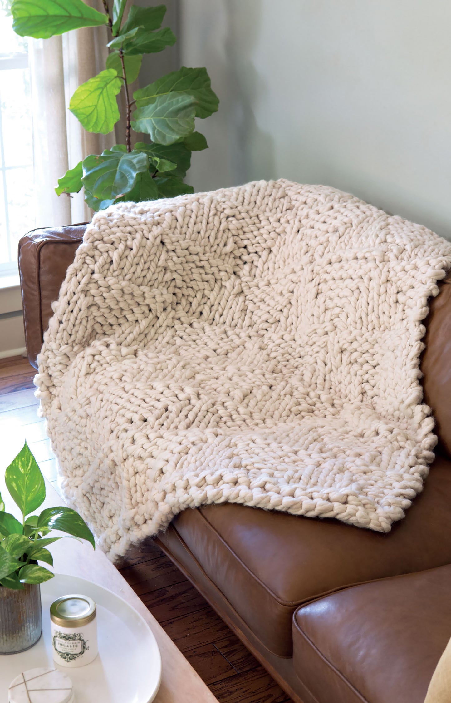 The Leisure Arts Oversized KNITS Digital Download features a chunky, cream-colored knit blanket made from jumbo yarn, shown draped over a brown leather sofa with green plants and sunlight nearby.