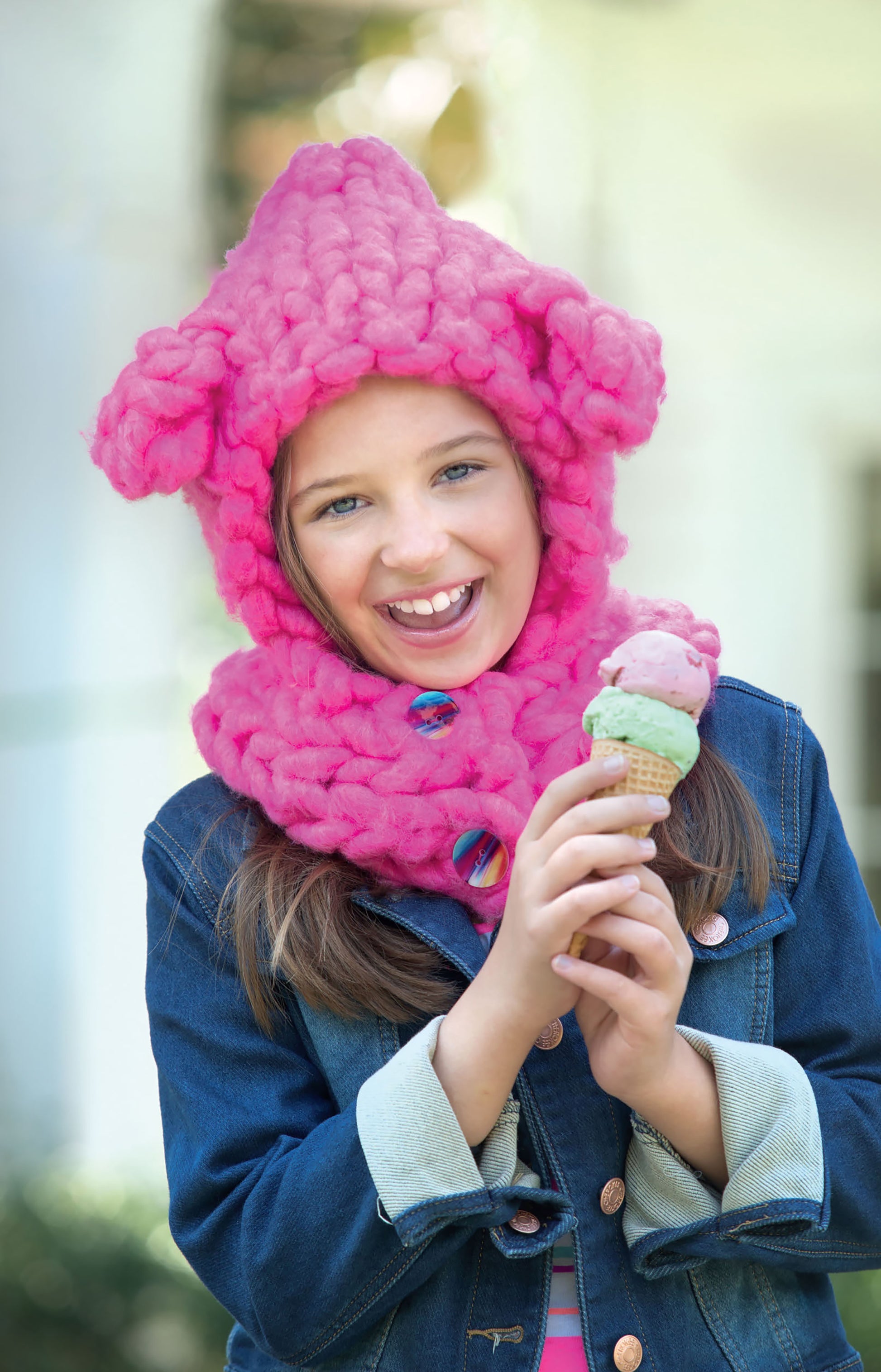 A happy child in a chunky, bright pink hat and scarf made from Oversized Knits by Leisure Arts enjoys a double-scoop ice cream cone. Dressed in a denim jacket outdoors, the child radiates joy.