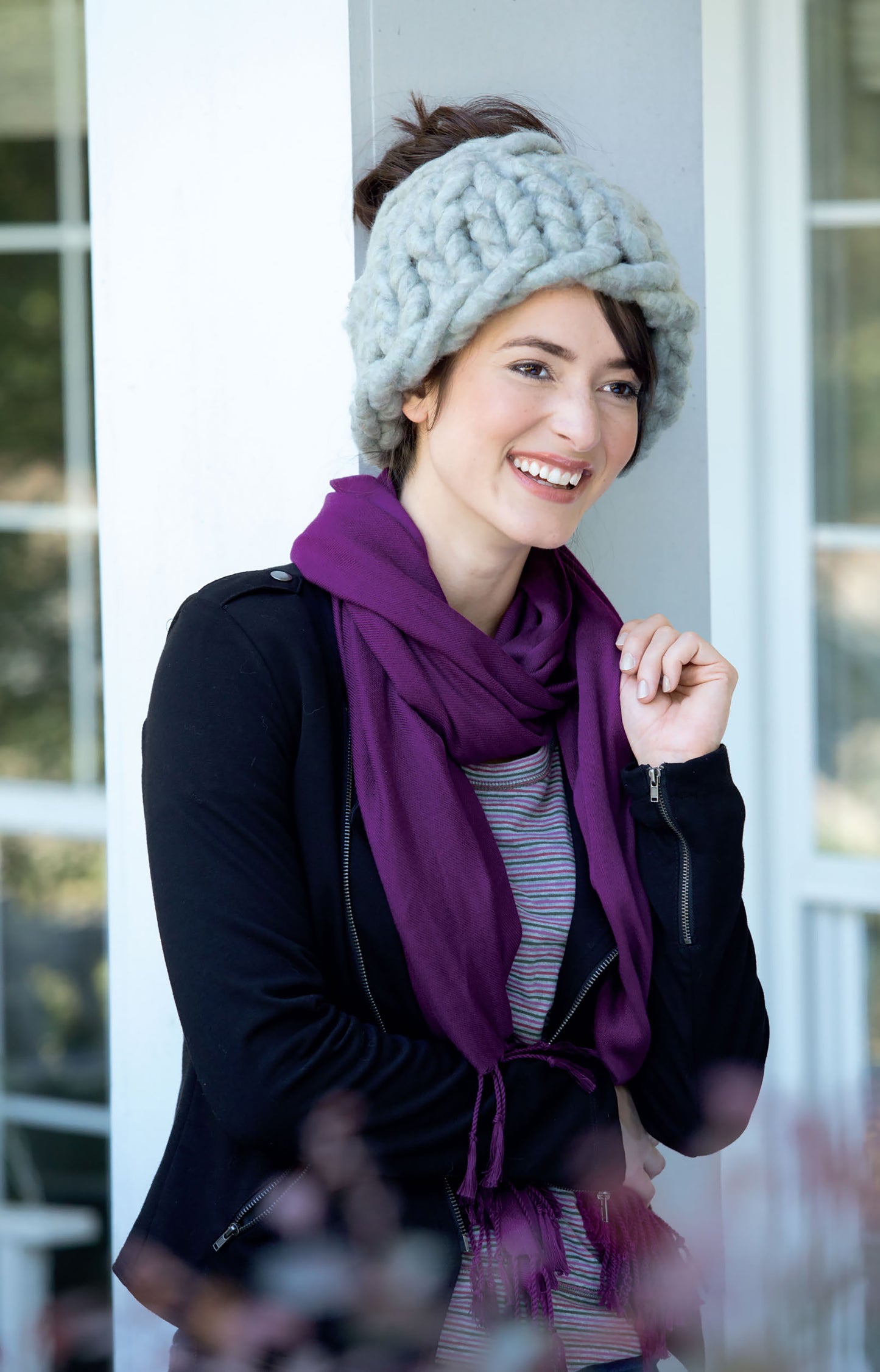 A smiling woman models an Oversized Knits chunky gray hat by Leisure Arts, paired with a purple scarf, black jacket, and striped top as she leans against a white wall in front of a window.