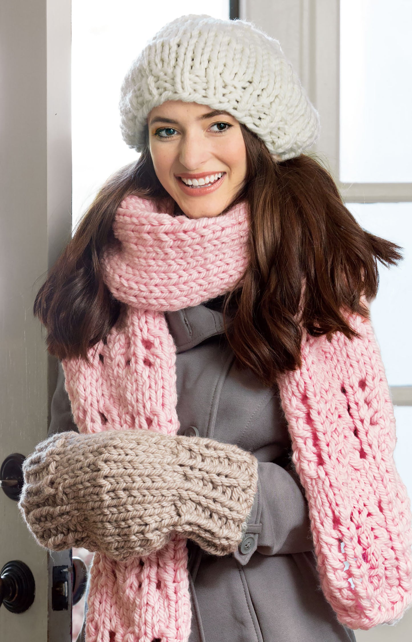 A woman in a chunky white hat, pink scarf, and beige mittens—created with Leisure Arts' Oversized Knits—stands indoors by a door, showcasing cozy patterns perfect for stylish home decor.