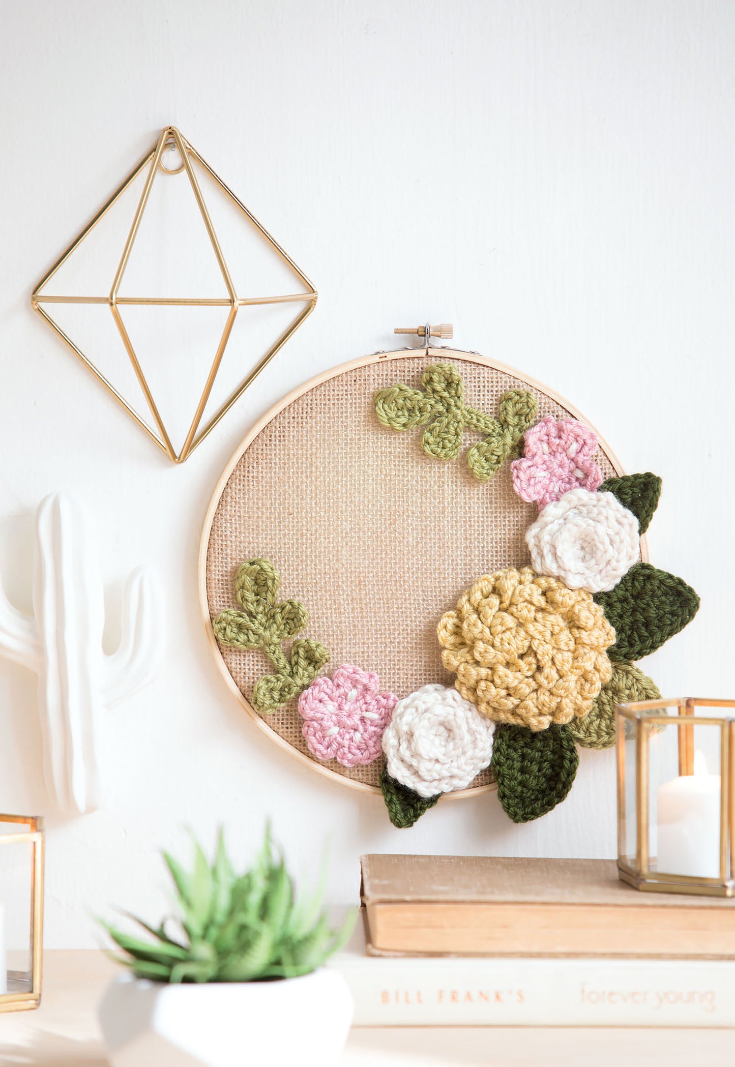Leisure Arts' "Quick Crochet With Flowers" is shown in a round hoop featuring floral and leaf designs, displayed on a wall near geometric gold decor, a white ceramic cactus, a green plant, books, and a glass candle holder.