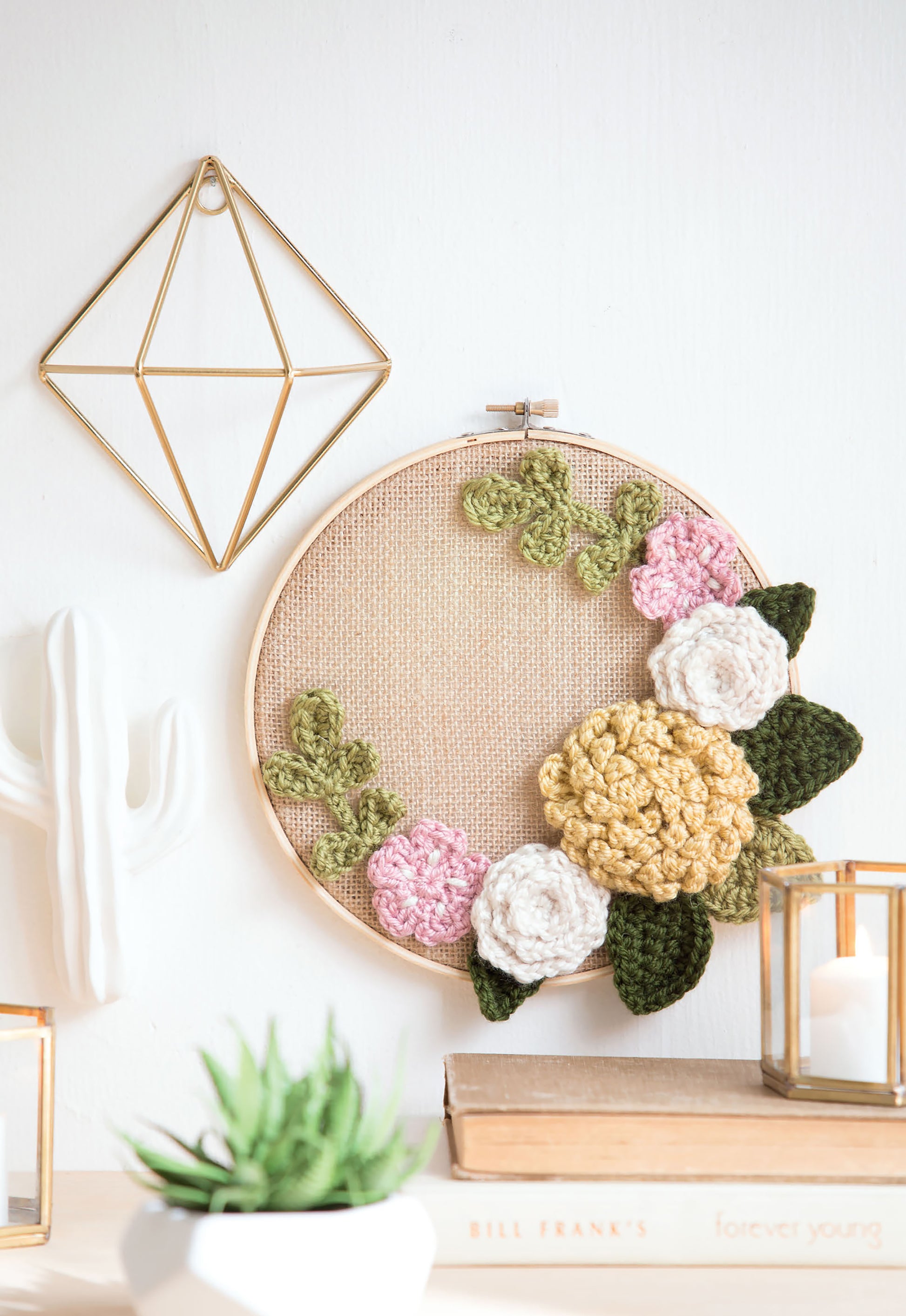 Leisure Arts' "Quick Crochet With Flowers" is shown in a round hoop featuring floral and leaf designs, displayed on a wall near geometric gold decor, a white ceramic cactus, a green plant, books, and a glass candle holder.