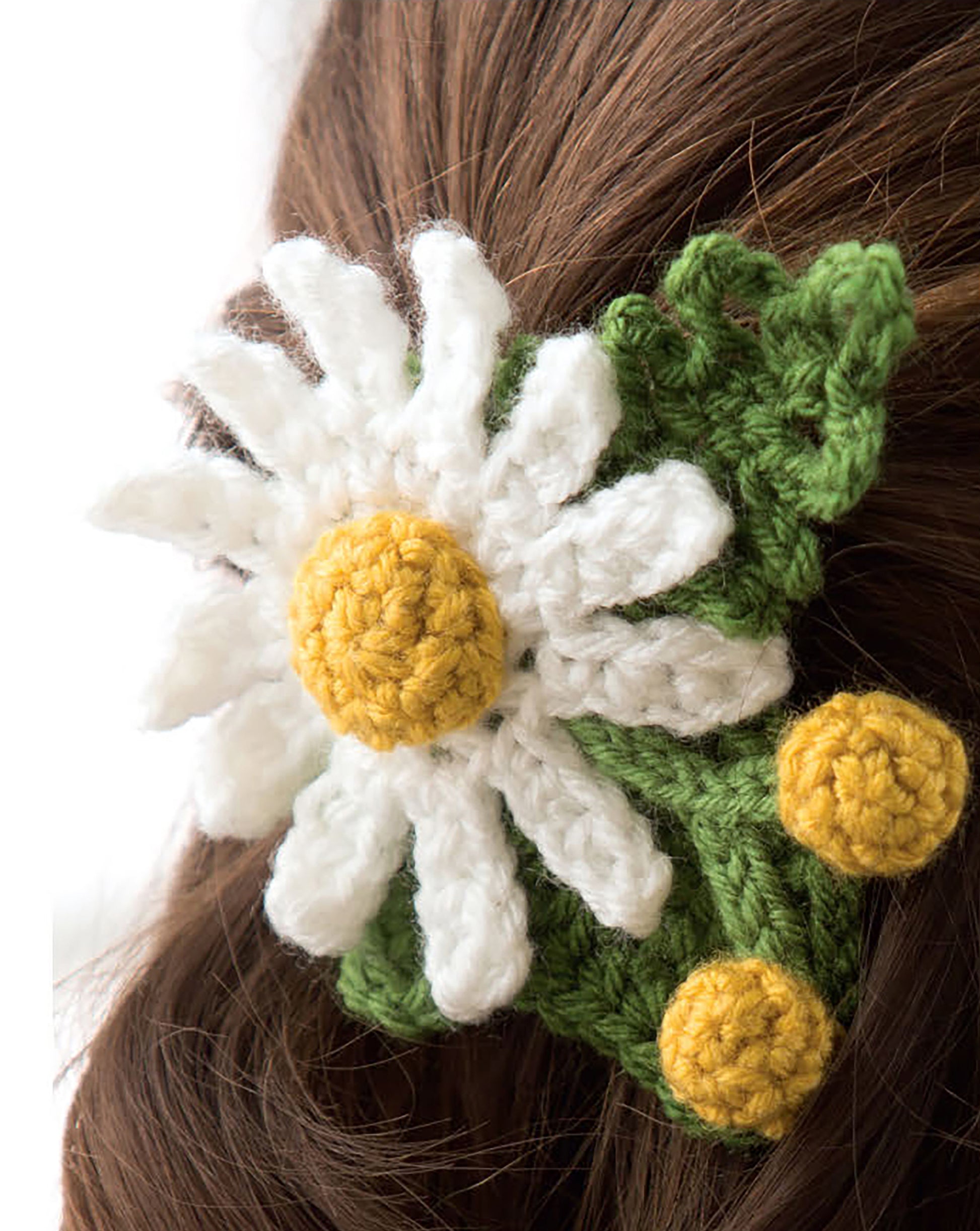 A crocheted hair accessory from Leisure Arts' "Quick Crochet With Flowers" features a white daisy with a yellow center, green leaves, and yellow buds, clipped into brown hair to display its lovely flower and leaf patterns.