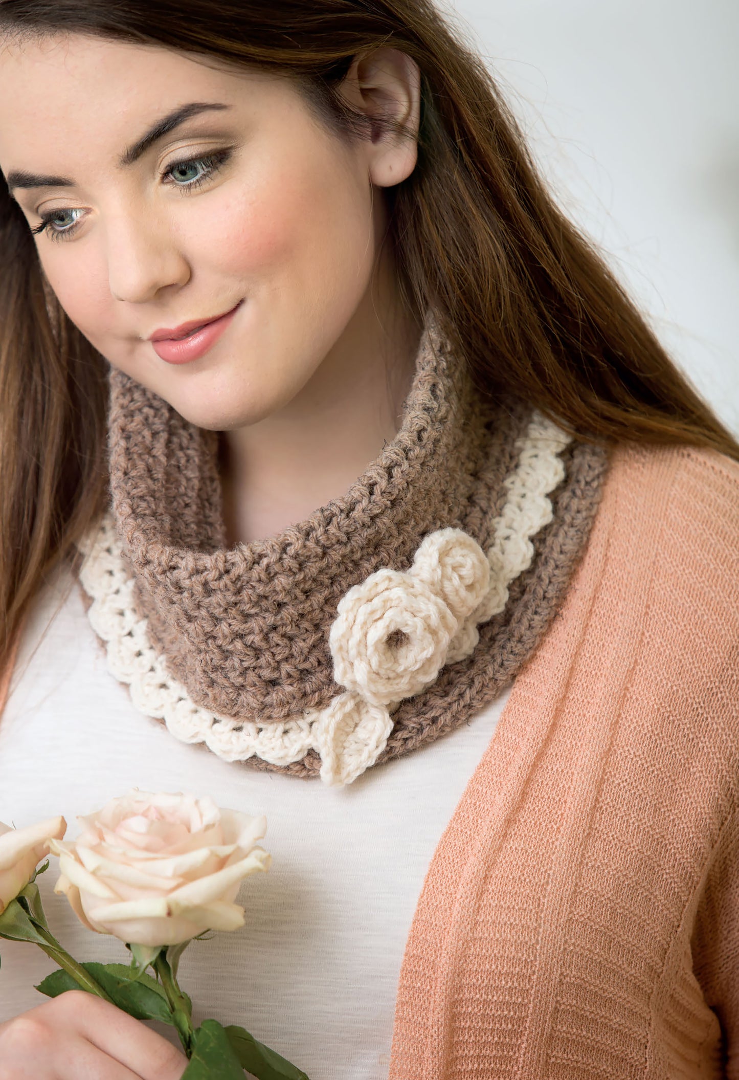A woman smiles softly in a peach cardigan and white top, wearing the "Quick Crochet With Flowers" cowl by Leisure Arts, crafted in brown and cream yarn with a decorative flower, while holding a pale pink rose.