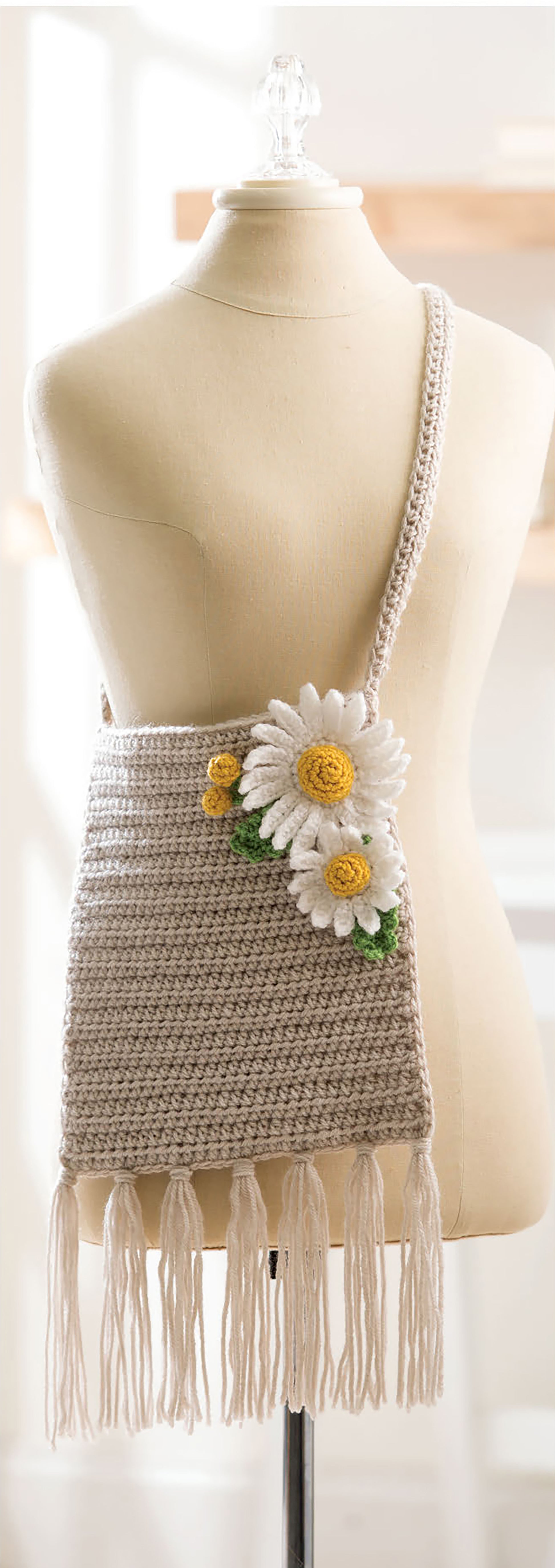 The "Quick Crochet With Flowers" bag by Leisure Arts is a beige, medium-weight yarn shoulder bag with long fringe and three crocheted white and yellow daisies, featuring subtle floral and leaf patterns. Displayed on a dress form.