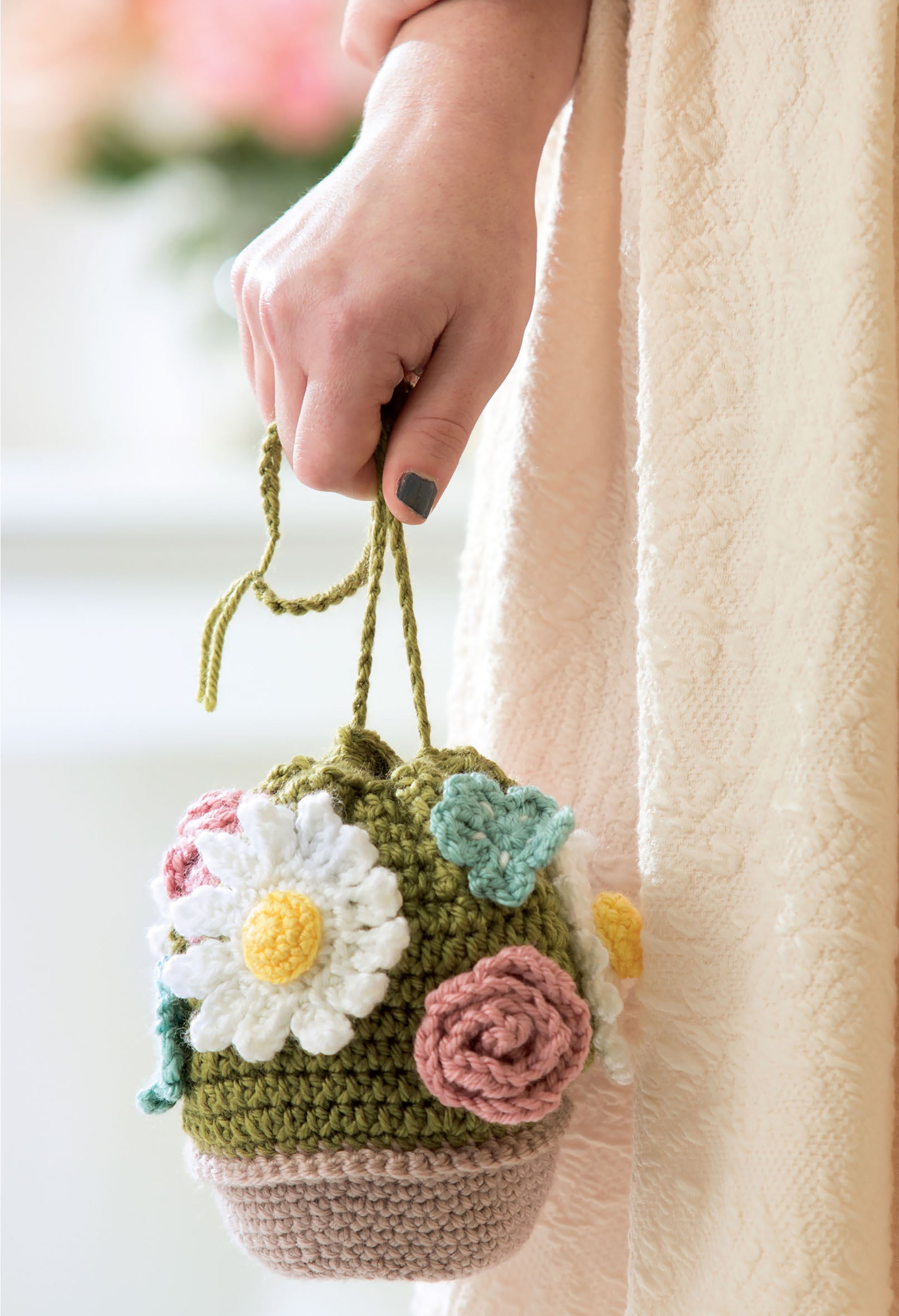A person in a cream dress holds a green "Quick Crochet With Flowers" bag by Leisure Arts, crafted with medium weight yarn, beige bottom, drawstring handle, and adorned with colorful crocheted flowers and leaves.