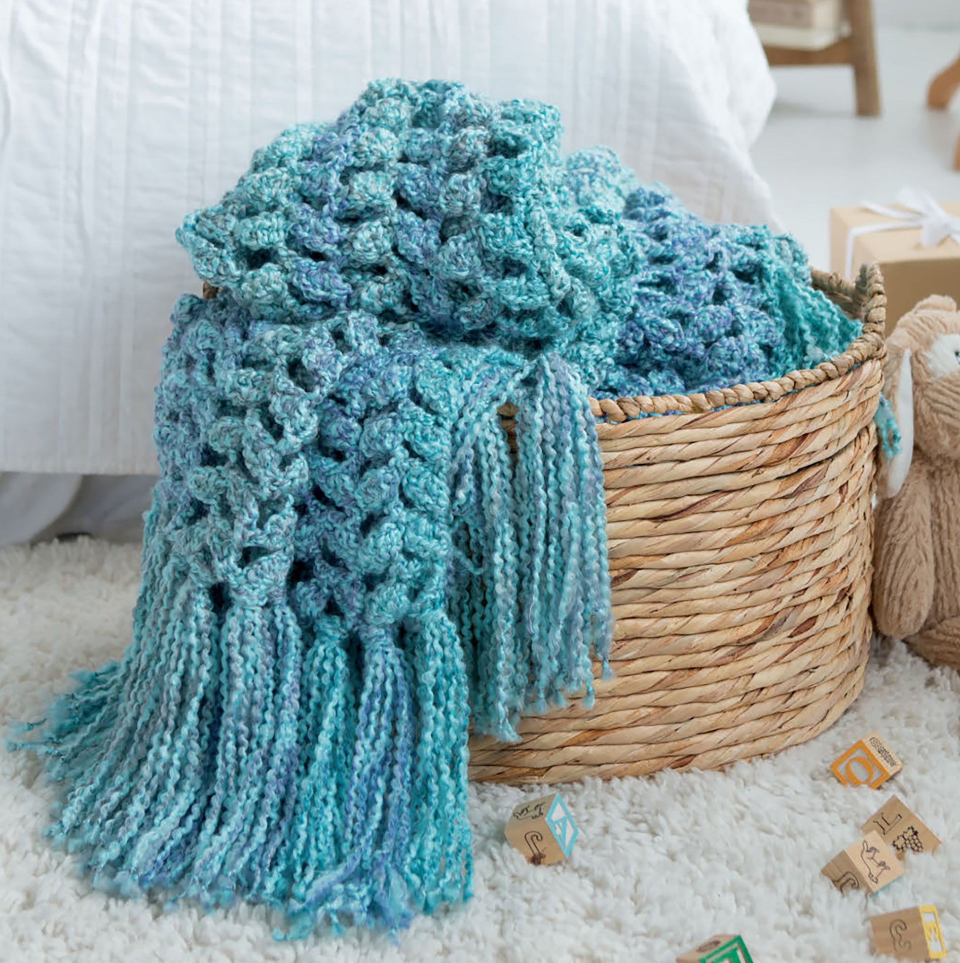 The Homespun Comfort Afghans by Leisure Arts, featuring a blue and teal crochet throw, is draped over a woven basket on a white carpet, with wooden letter blocks nearby in a bright and inviting room.