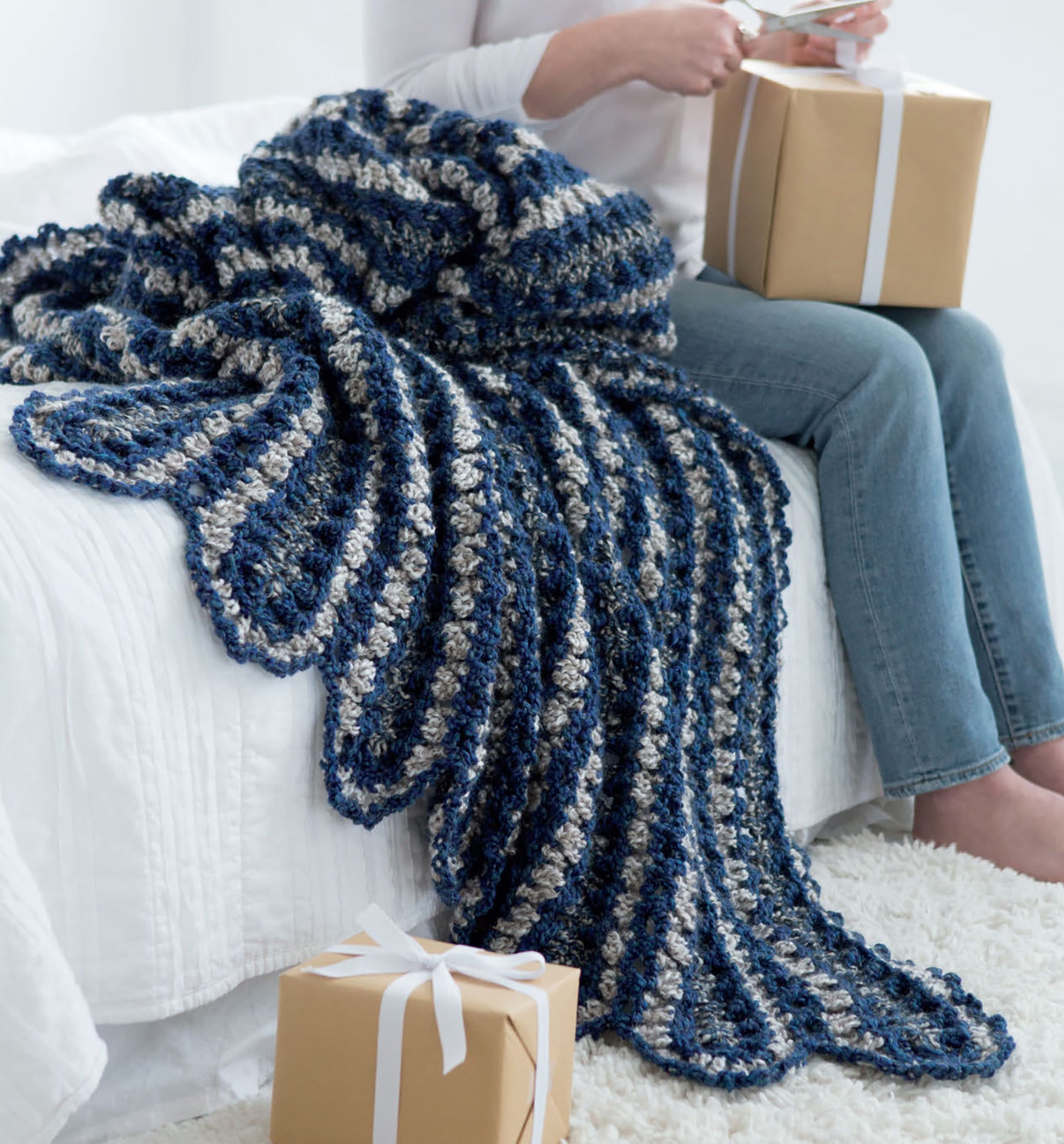 A person on a bed holds a wrapped gift and card, with the Leisure Arts Homespun Comfort Afghans draped over the bed. Another wrapped gift rests on a white rug nearby.