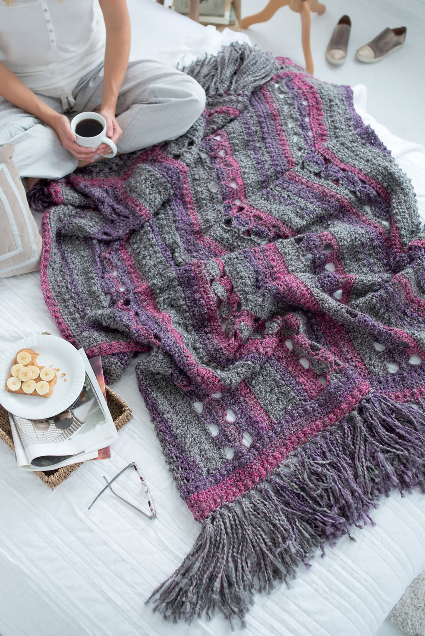 A person sits cross-legged on a bed, sipping coffee beside a gray and purple blanket made with Leisure Arts' Homespun Comfort Afghans—a cozy crochet throw. Sliced bananas, magazines, glasses, and slippers lie nearby on the floor.