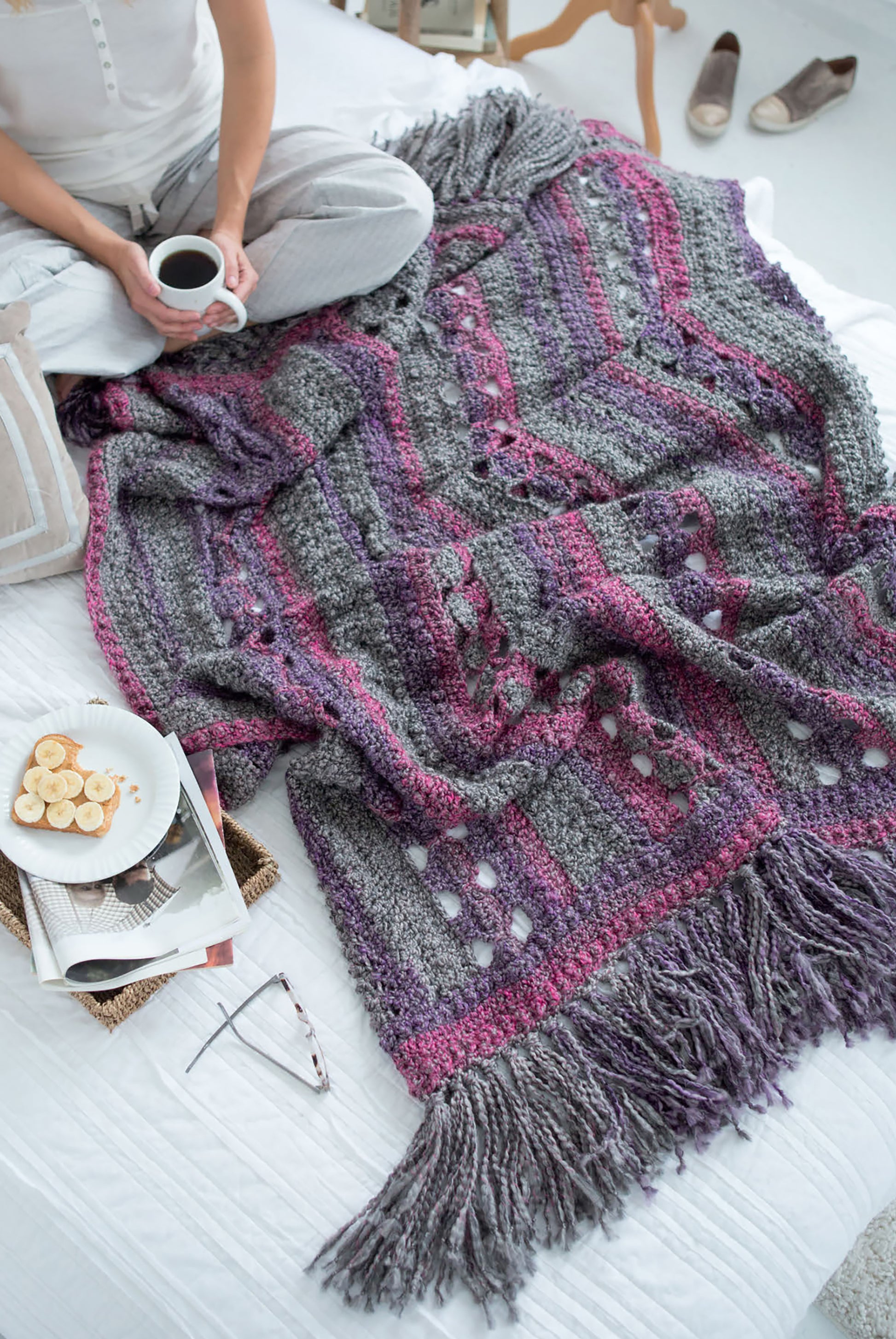 A person sits cross-legged on a bed, sipping coffee beside a gray and purple blanket made with Leisure Arts' Homespun Comfort Afghans—a cozy crochet throw. Sliced bananas, magazines, glasses, and slippers lie nearby on the floor.