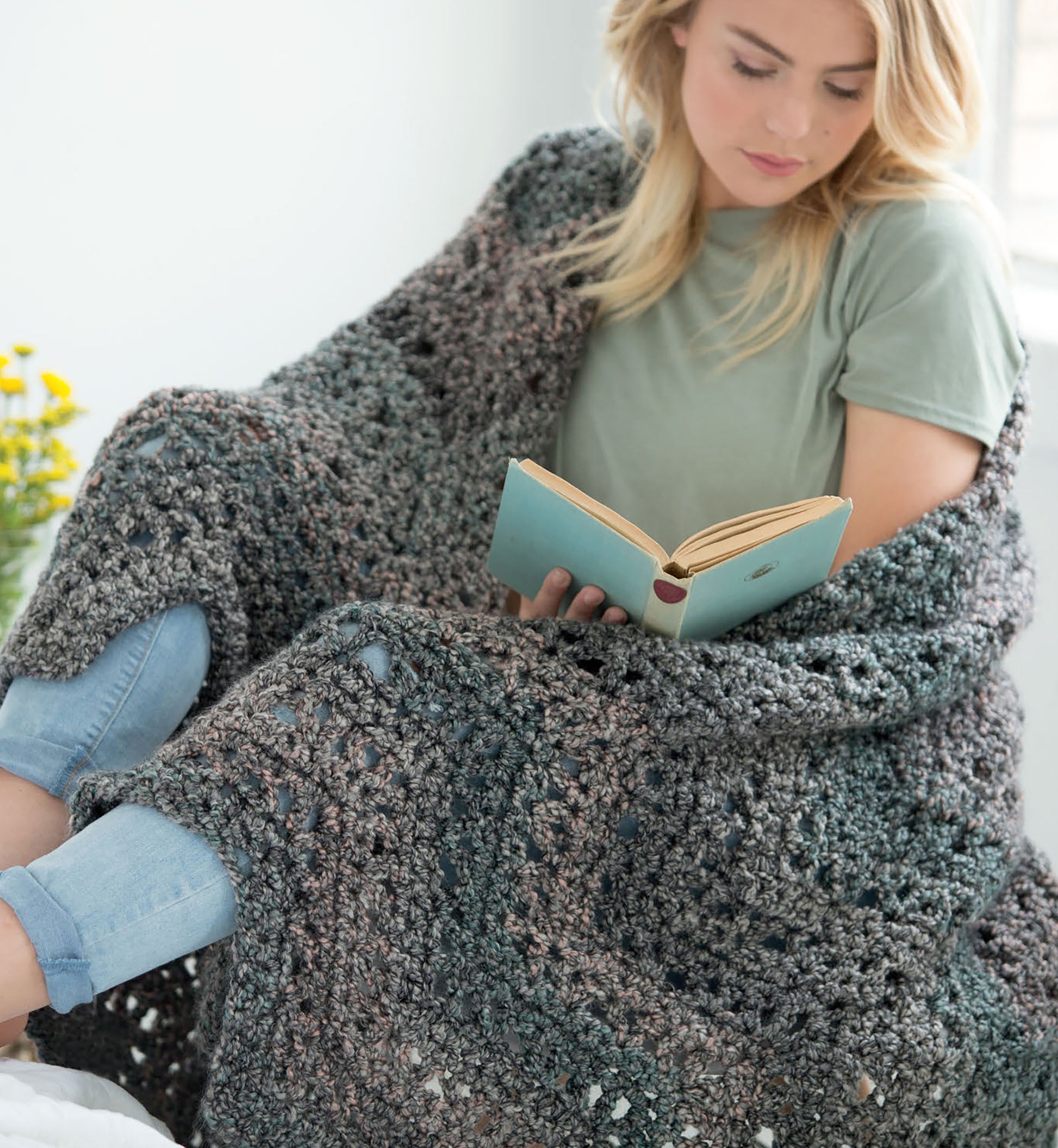 Wrapped in a chunky gray knit blanket from the Homespun Comfort Afghans by Leisure Arts, a blonde woman in a green t-shirt and jeans reads, inspired by cozy crochet throw patterns with a vase of yellow flowers nearby.