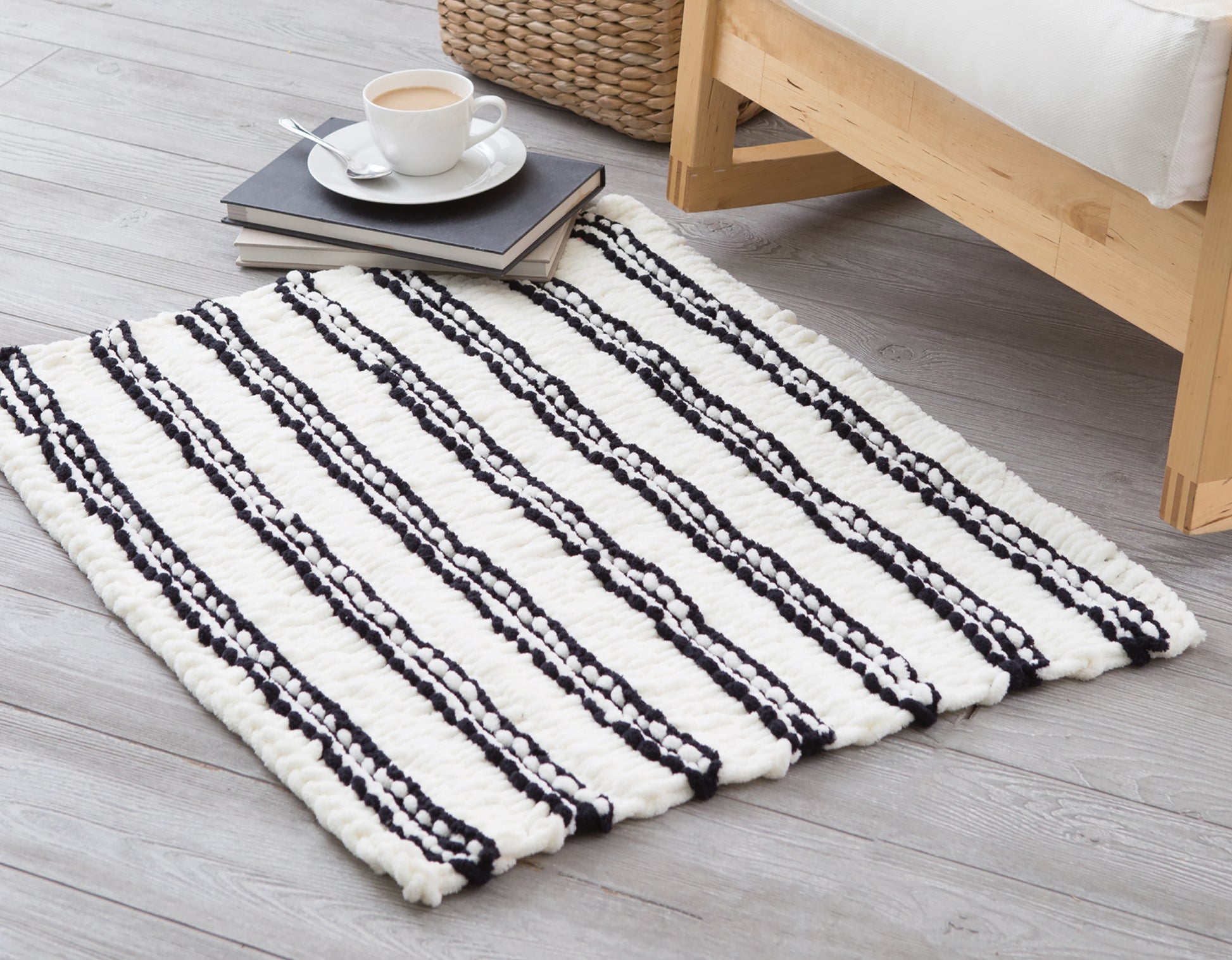 The Leisure Arts "Loops & Twists—Knitting Without Needles" white woven rug with black stripes rests on a light wooden floor by a bed. Beside it, coffee sits atop books, while a wicker basket appears in the background.