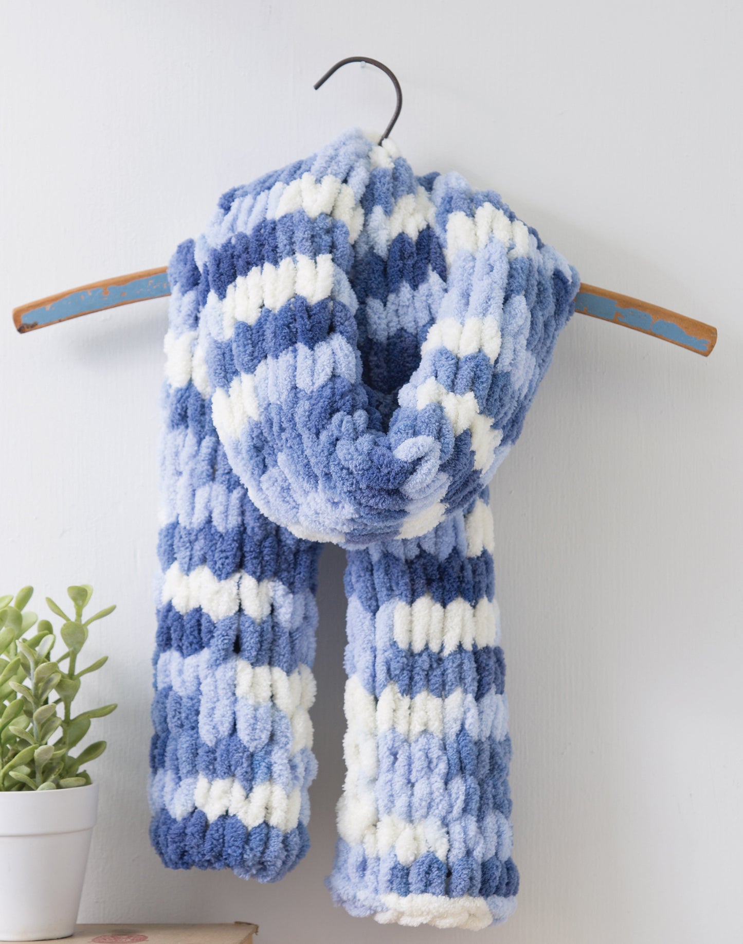 A soft, chunky striped scarf in blue, white, and light blue—made with Loops & Twists—Knitting Without Needles by Leisure Arts—hangs on a blue wooden hanger against a white wall. Great for beginner projects. A small potted plant sits nearby.