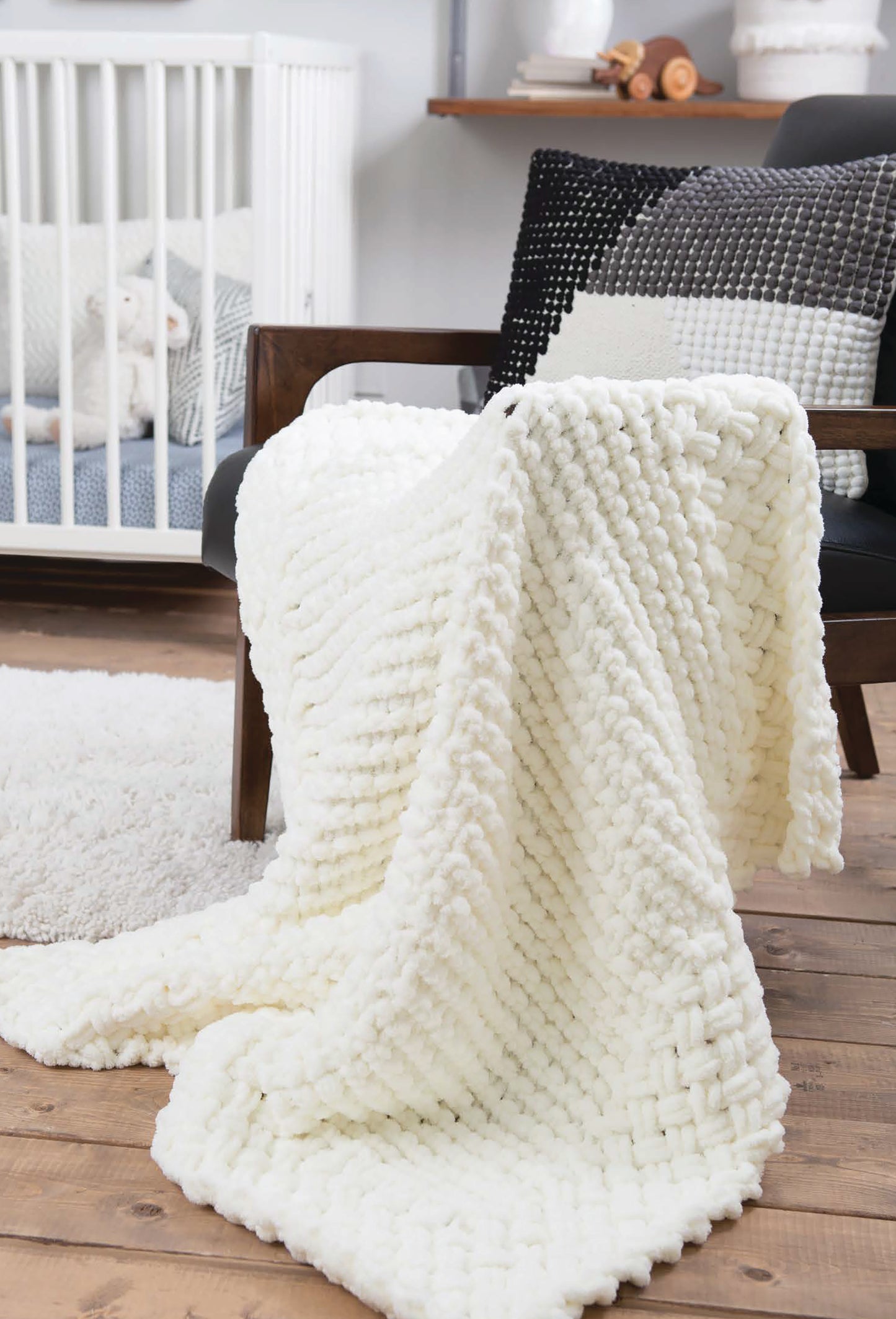 A Leisure Arts Baby Loops & Twists soft, chunky white knit blanket, made with loop yarn and no needles, is draped over a black armchair with textured pillows. In back, a white crib with a stuffed animal and light rug sit on wood floors.
