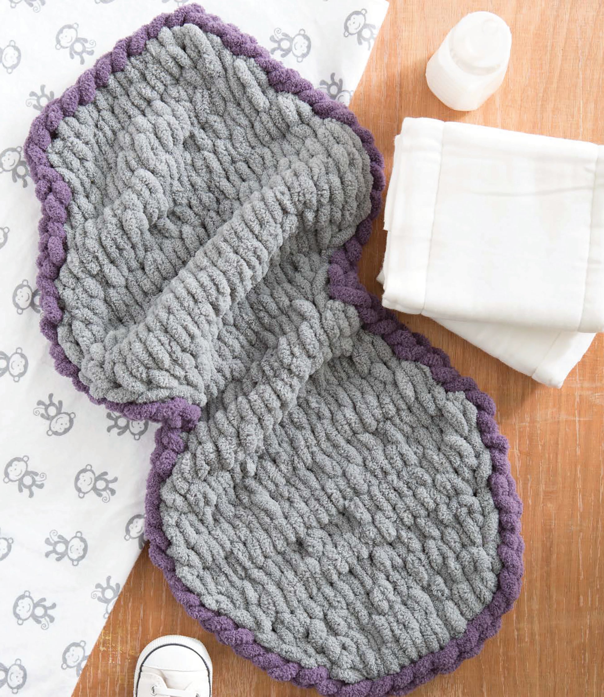 The Baby Loops & Twists—No-Needle Knitting for Baby! by Leisure Arts is a gray and purple loop yarn wrap on wood, beside a white blanket, baby bottle, sheep-patterned cloth, and baby shoe—ideal for beginner knitters.