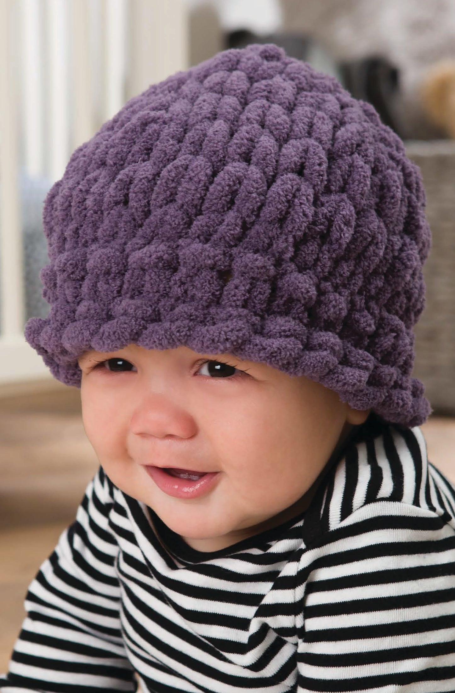 A smiling baby wears a chunky purple knit hat made with Baby Loops & Twists—No-Needle Knitting for Baby! by Leisure Arts, plus a black-and-white striped shirt, sitting indoors on a wooden floor with a blurred background.