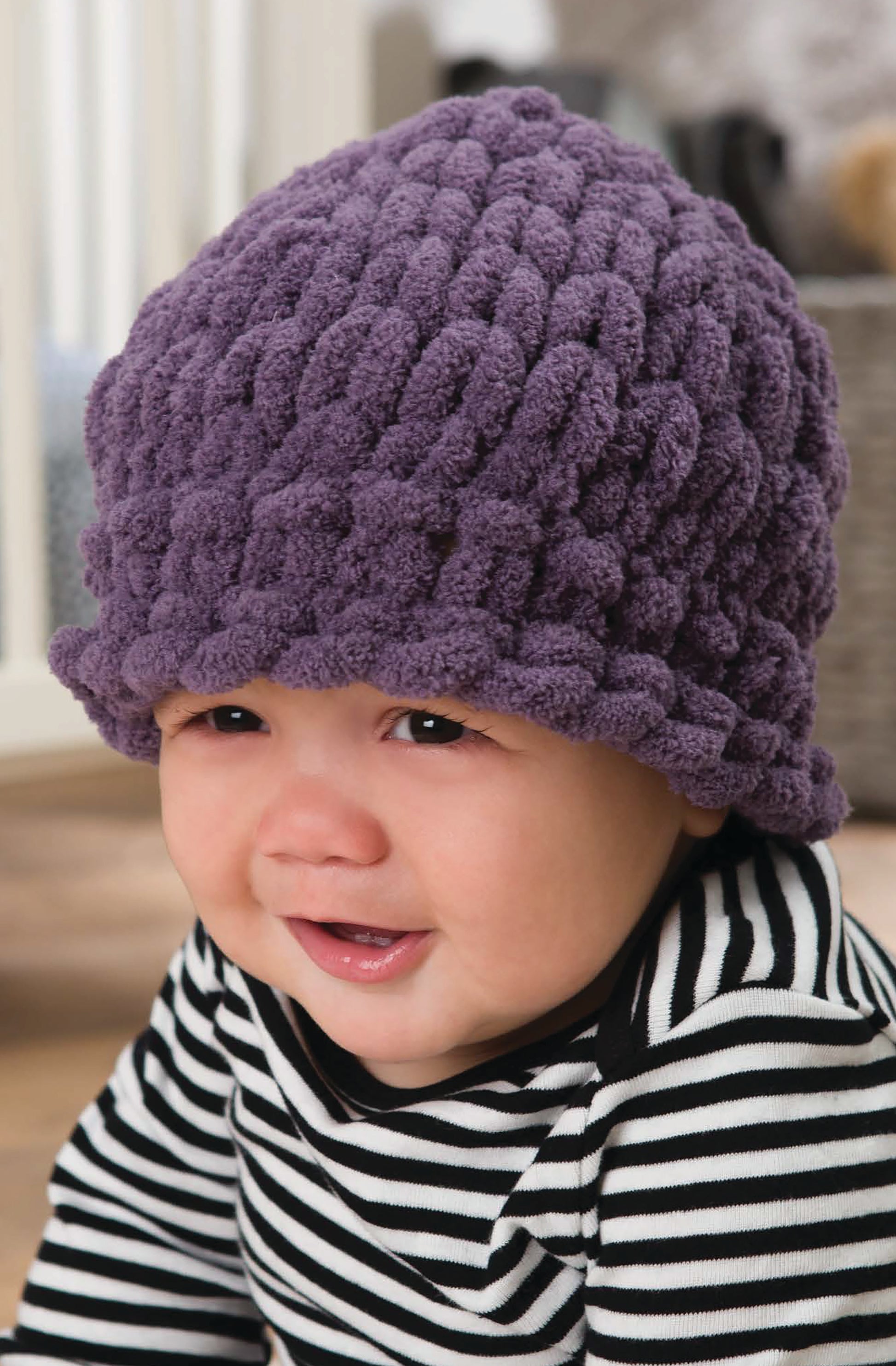 A smiling baby wears a chunky purple knit hat made with Baby Loops & Twists—No-Needle Knitting for Baby! by Leisure Arts, plus a black-and-white striped shirt, sitting indoors on a wooden floor with a blurred background.