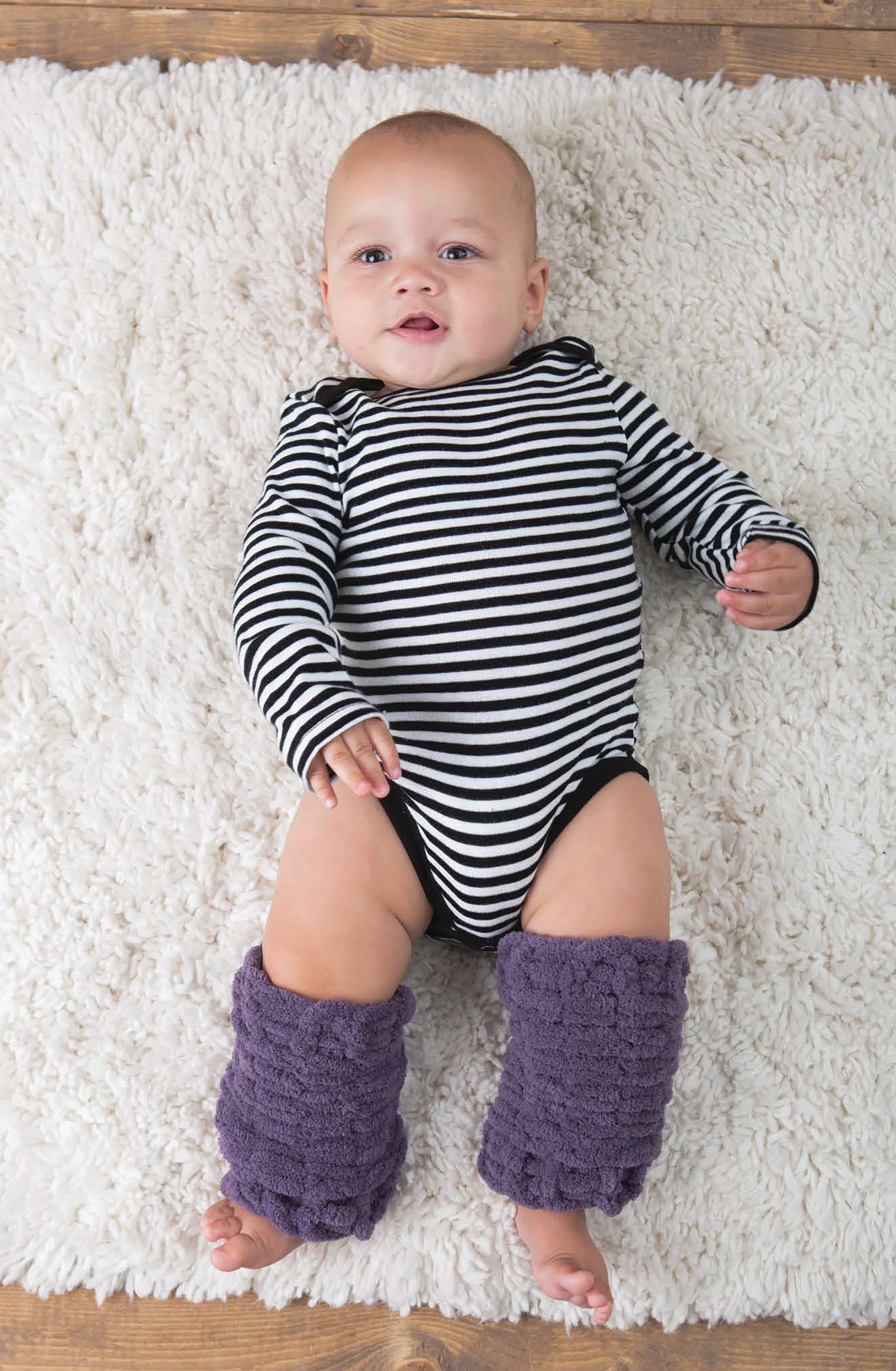 A baby in handmade purple leg warmers from Leisure Arts' "Baby Loops & Twists—No-Needle Knitting for Baby!" wears a striped onesie and lies on a white fluffy rug, gazing up with a small smile.