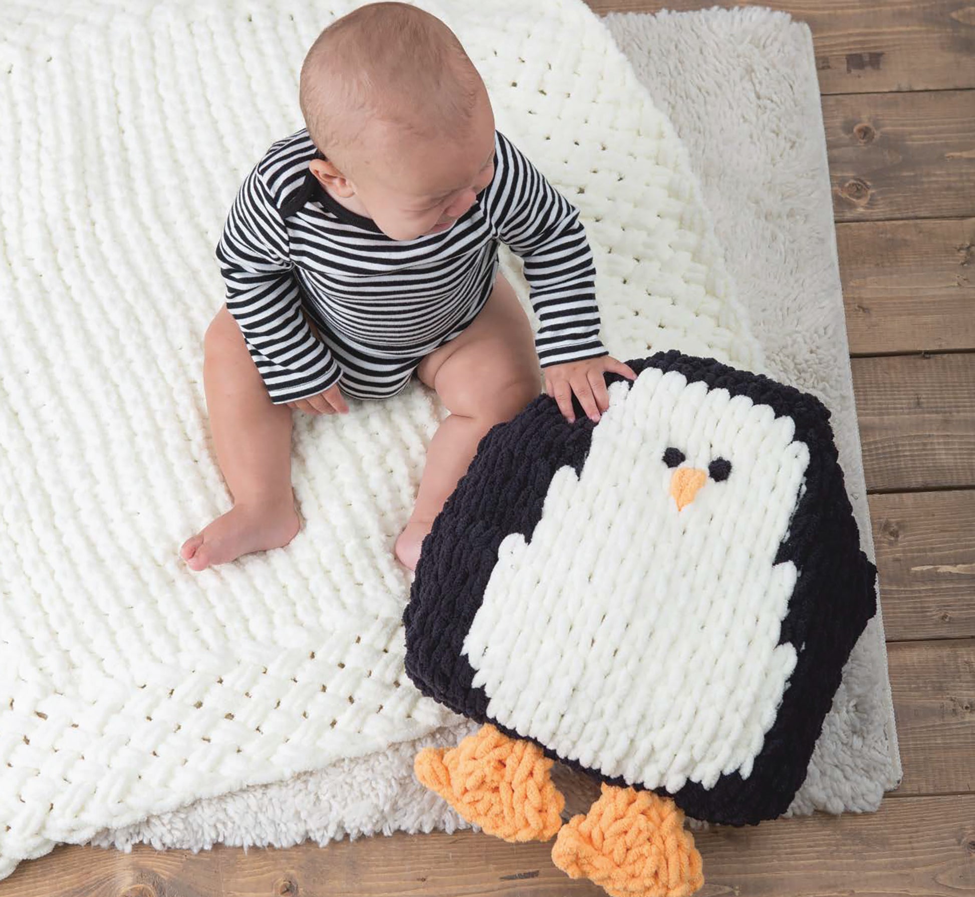 A baby in a black-and-white striped onesie sits on a soft white blanket, reaching for a plush penguin made with Baby Loops & Twists—No-Needle Knitting for Baby! by Leisure Arts, its orange feet resting on the wooden floor.