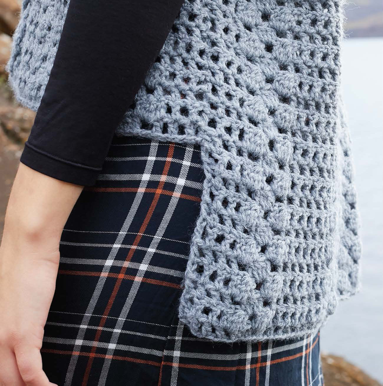 A person wears the Leisure Arts Northern Lights Crochet vest (digital download), Icelandic-inspired in light blue, layered over a black long-sleeve shirt and navy checkered skirt with orange and white lines near waterfront outdoors.