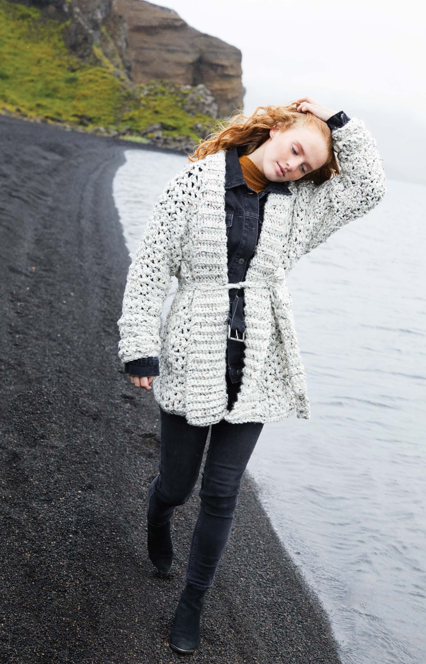 A woman with red hair, in black jeans and boots, walks on a black sand beach wearing a chunky cardigan made from Leisure Arts’ Northern Lights Crochet Digital Download pattern on a cloudy day.