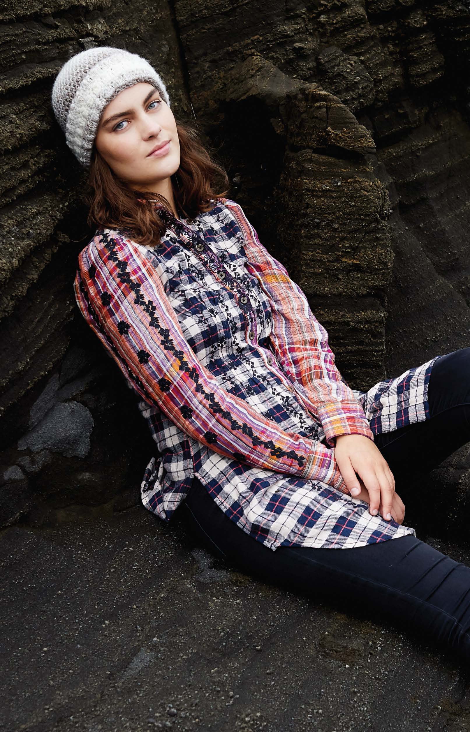 A young woman wearing a beanie made from the Northern Lights Crochet Digital Download by Leisure Arts, along with a colorful plaid and floral shirt and dark pants, sits against a textured rock wall, gazing calmly at the camera.