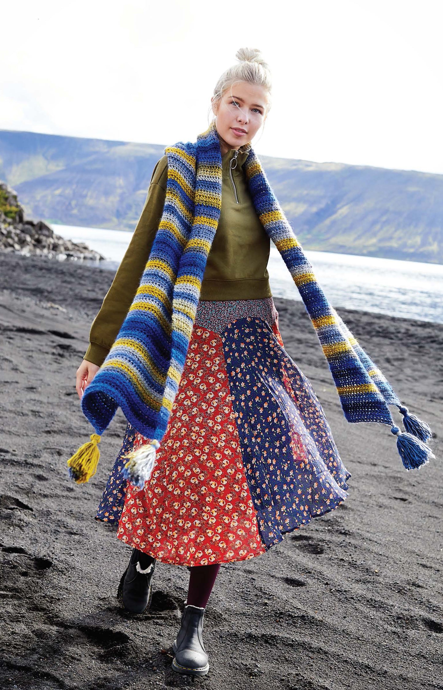 A woman with blond hair in a bun wears a green sweater, long colorful patchwork skirt, and a striped scarf made from the Northern Lights Crochet Digital Download by Leisure Arts, walking on a black sand beach with cliffs and a cloudy sky in the background.