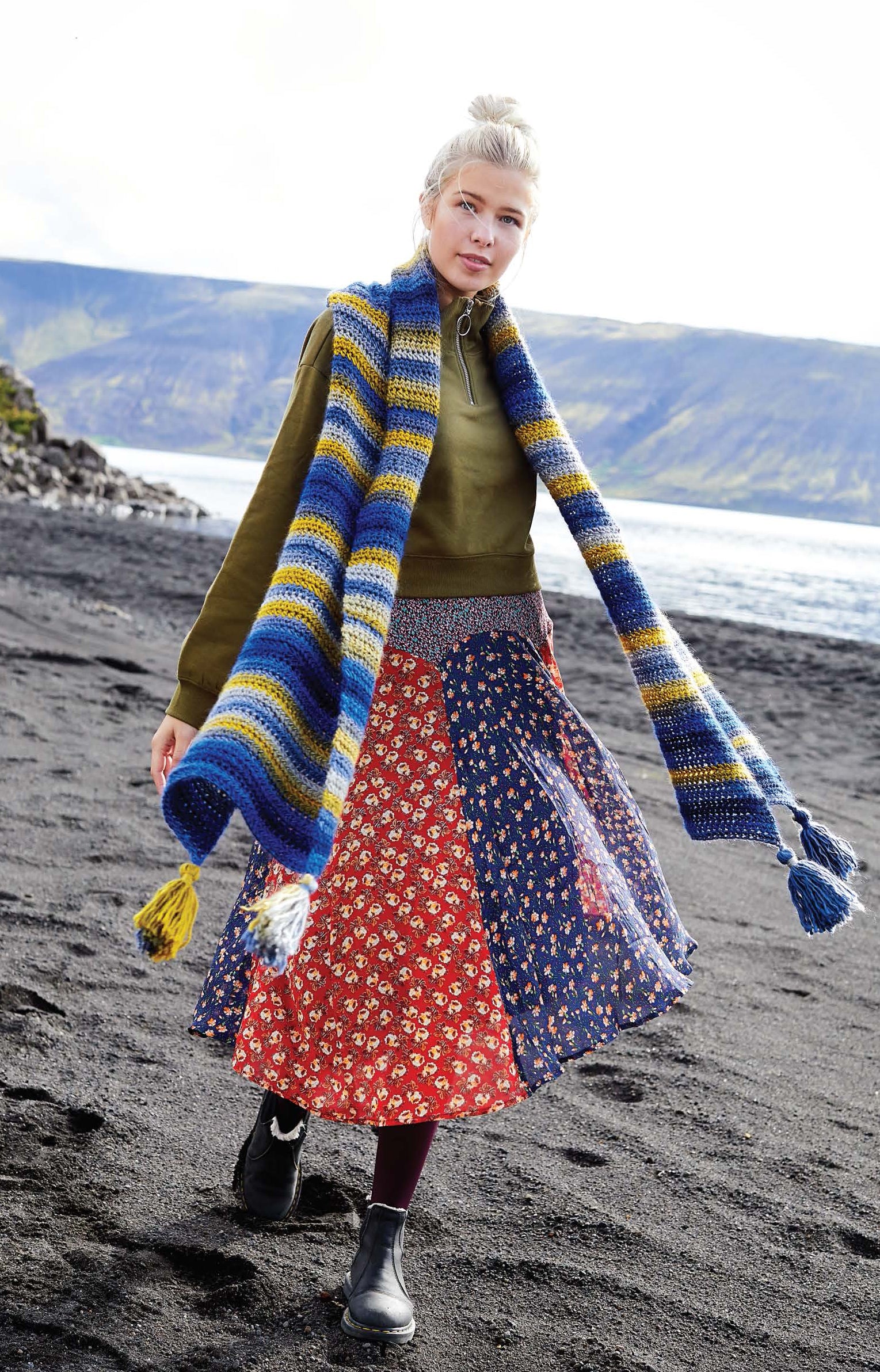 A woman with blond hair in a bun wears a green sweater, long colorful patchwork skirt, and a striped scarf made from the Northern Lights Crochet Digital Download by Leisure Arts, walking on a black sand beach with cliffs and a cloudy sky in the background.