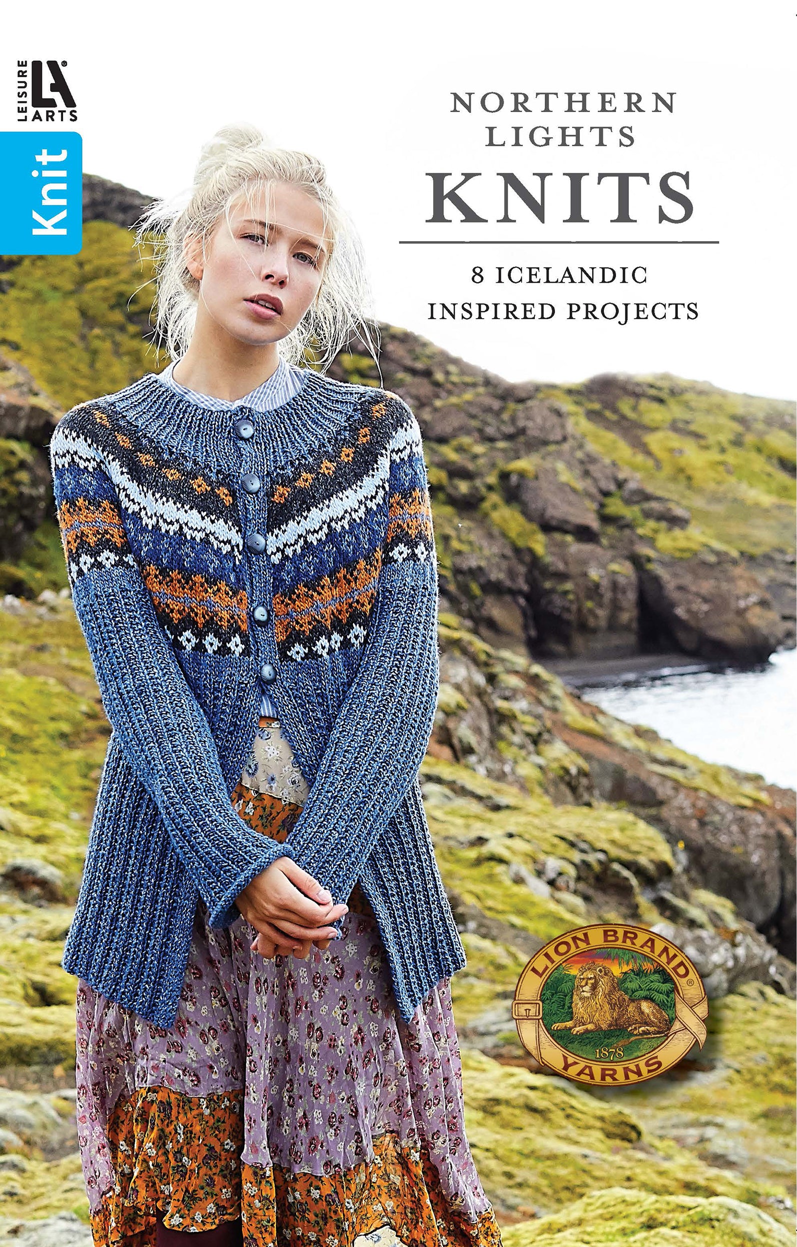 A woman stands on a rocky, grassy coastline wearing a blue, patterned knit cardigan over a floral dress. Text reads: Northern Lights Knits by Leisure Arts—8 Icelandic inspired knit sweaters and easy knitting patterns.