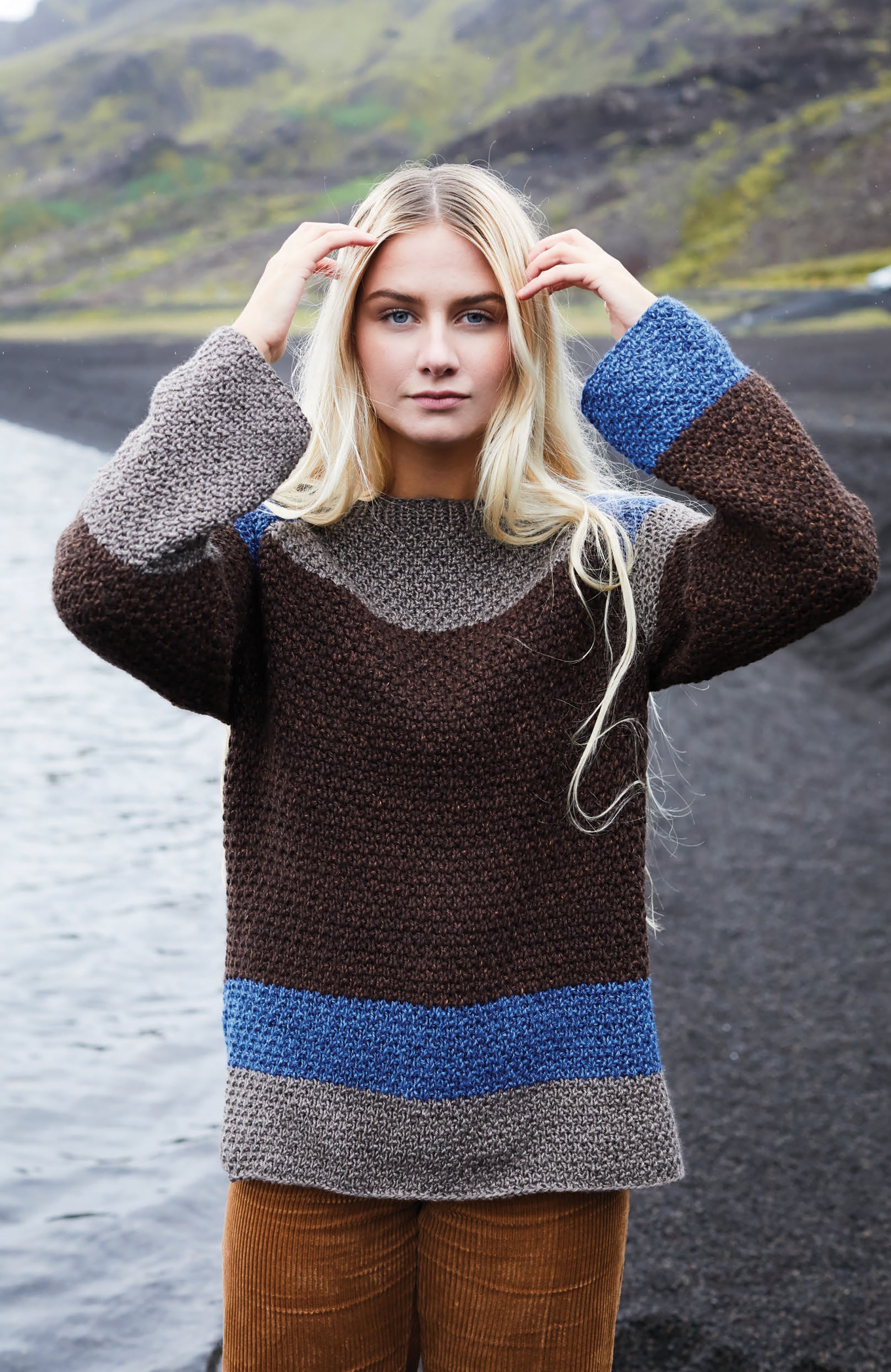 A young woman with long blonde hair stands by a rocky shoreline, wearing the Northern Lights Knits chunky sweater in brown, blue, and gray by Leisure Arts, with one hand touching her head and a neutral expression.