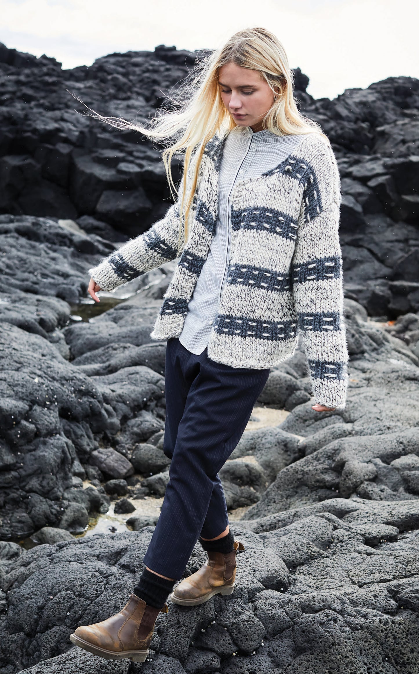 A person with long blonde hair walks on black volcanic rocks, wearing a thick, patterned Northern Lights Knits sweater by Leisure Arts over a light shirt with dark pants and brown boots, wind blowing through their hair.