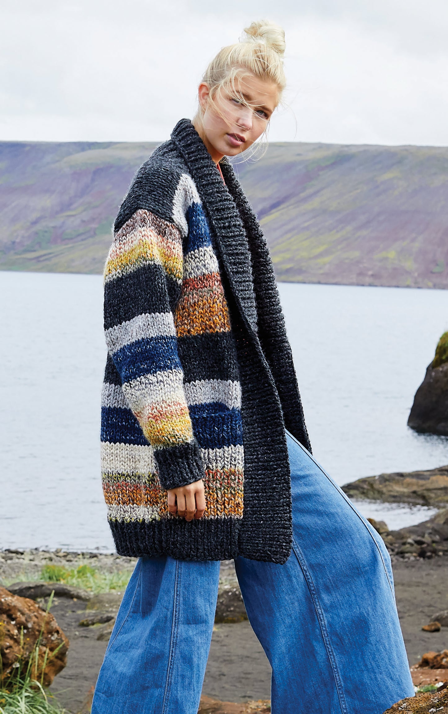 A person stands outdoors by water and mountains, wearing the Northern Lights Knits chunky sweater from Leisure Arts and wide-leg blue jeans, with cloudy skies enhancing the serene, natural setting.
