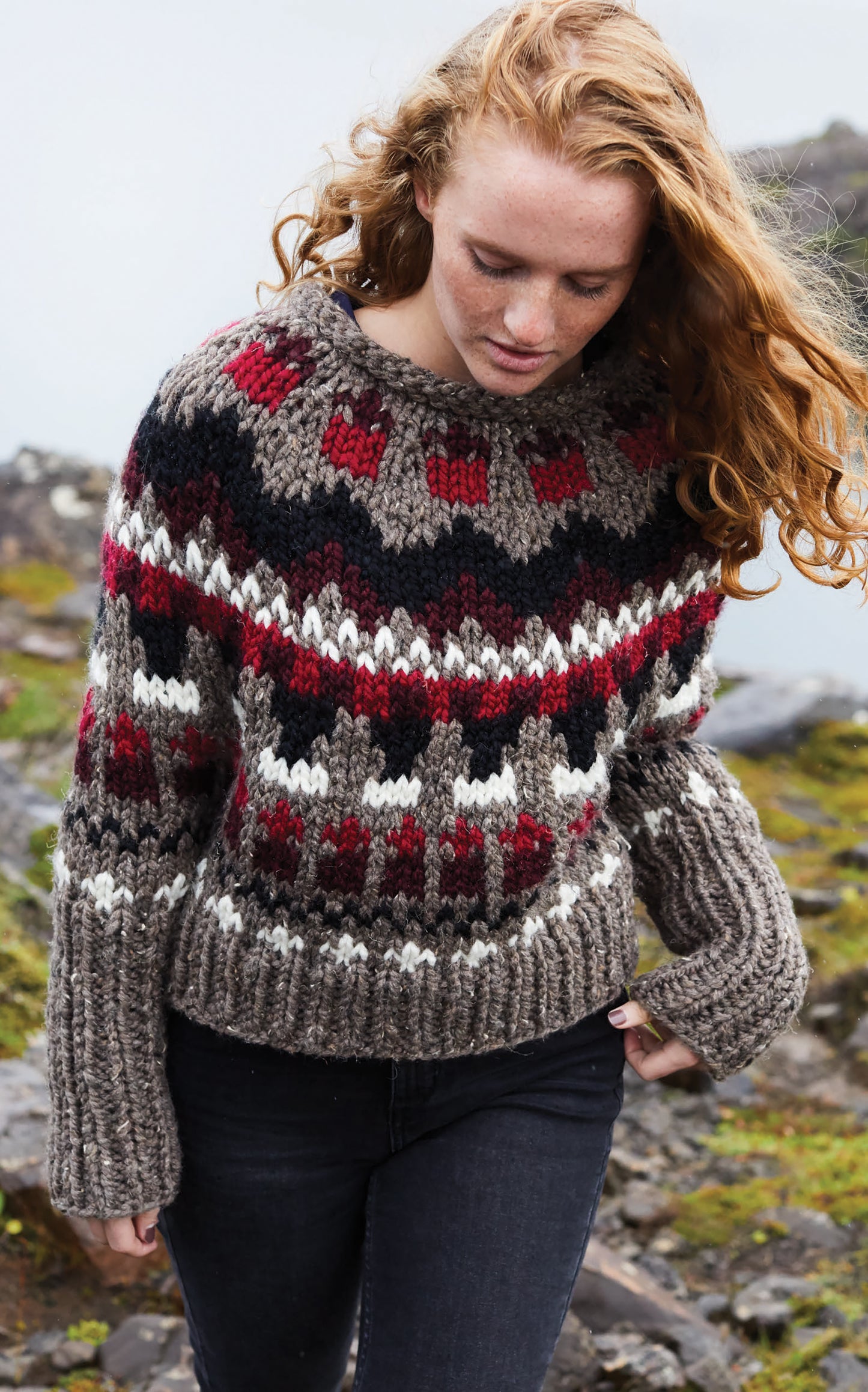 A person with long red hair stands outdoors near rocks and moss, wearing Leisure Arts' Northern Lights Knits sweater in brown, red, white, and black wool yarn as the wind blows their hair.