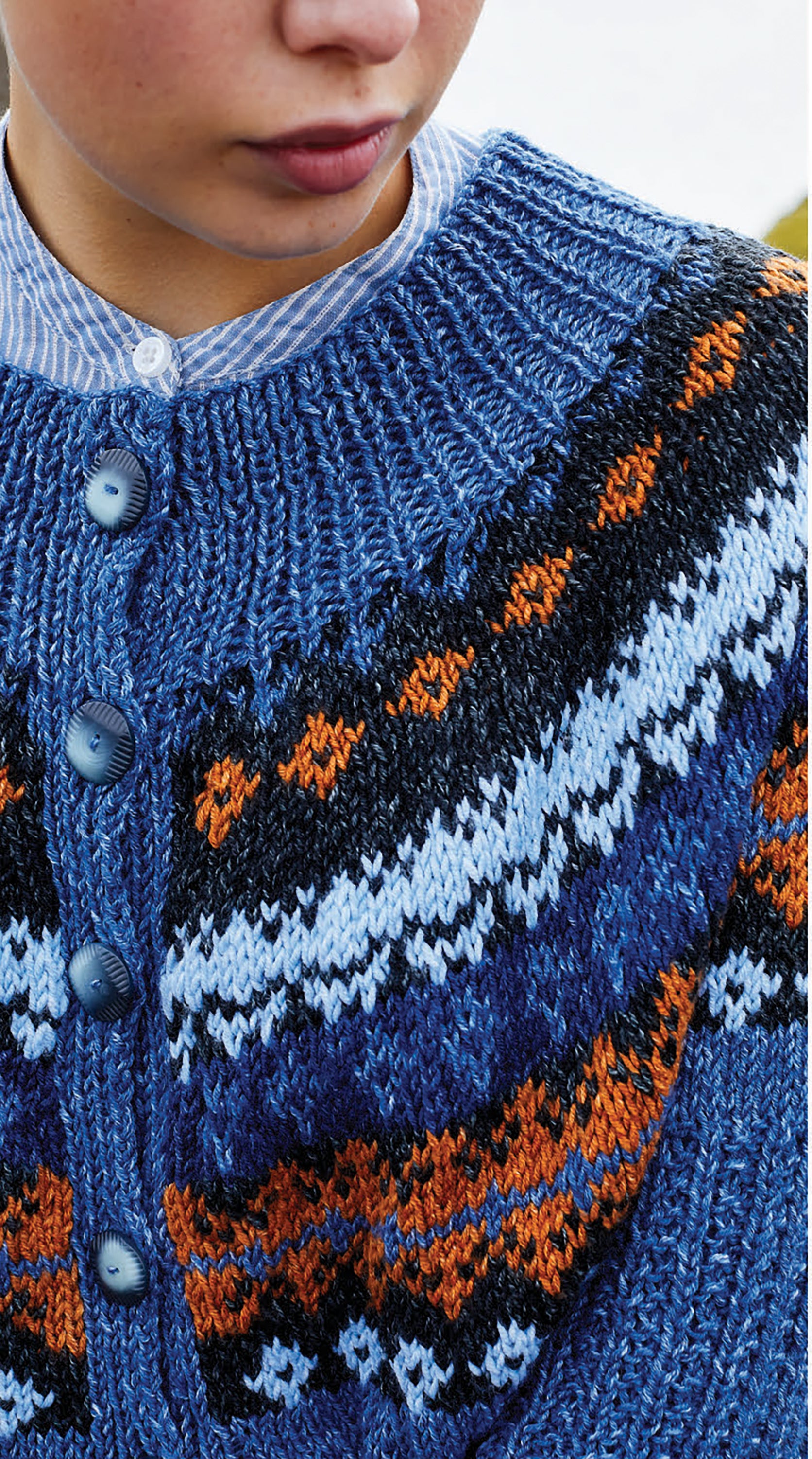 Wearing the Northern Lights Knits sweater by Leisure Arts, featuring a blue and orange pattern with large blue buttons, layered over a blue and white striped shirt; only the lower face and upper torso are shown.