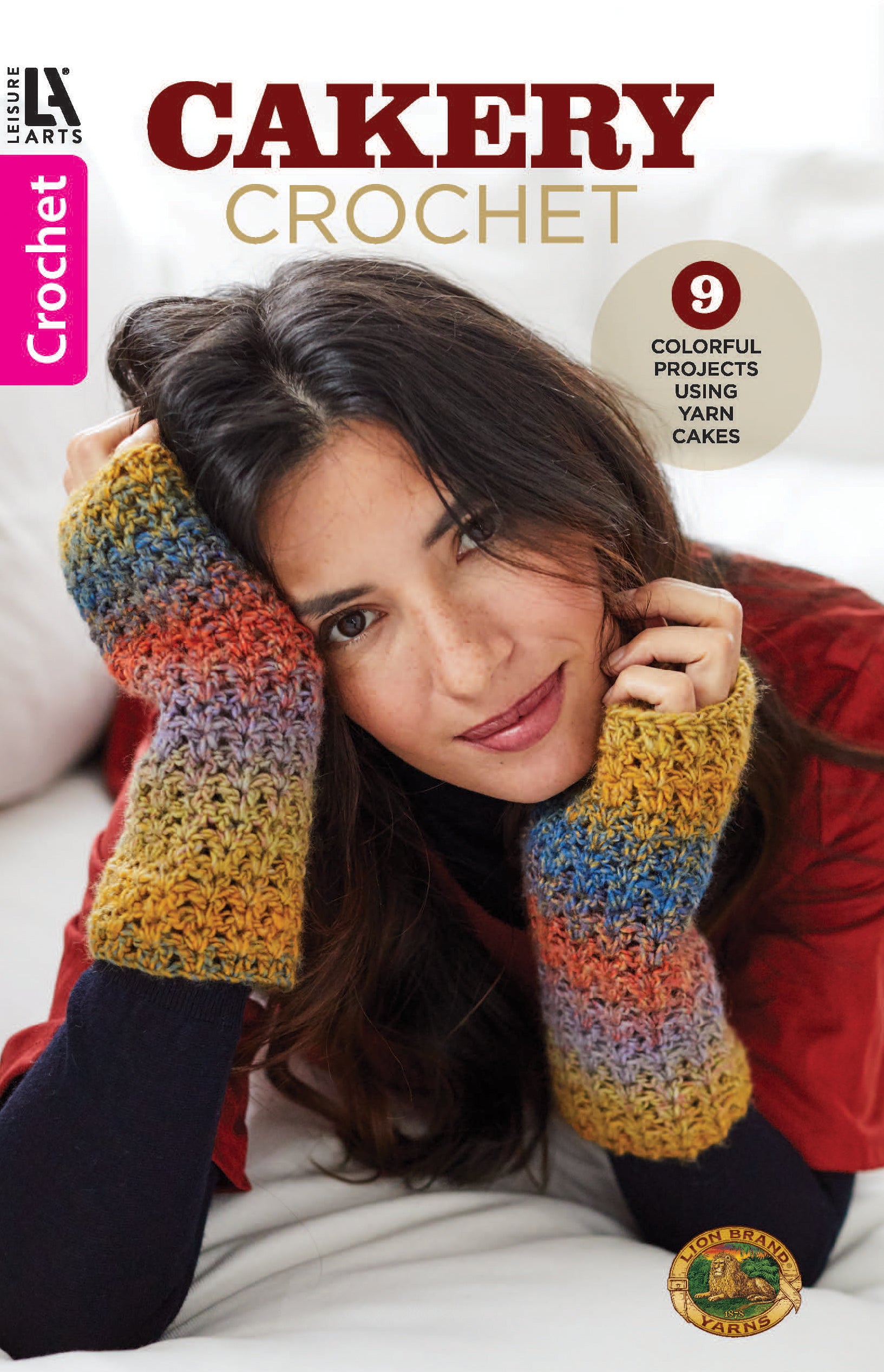 A woman with long dark hair wears colorful crocheted fingerless gloves, resting her face on her hands. Text reads: “Cakery Crochet by Leisure Arts – 9 Colorful Projects Made with Lion Brand Yarn Cakes and step-by-step instructions.”.