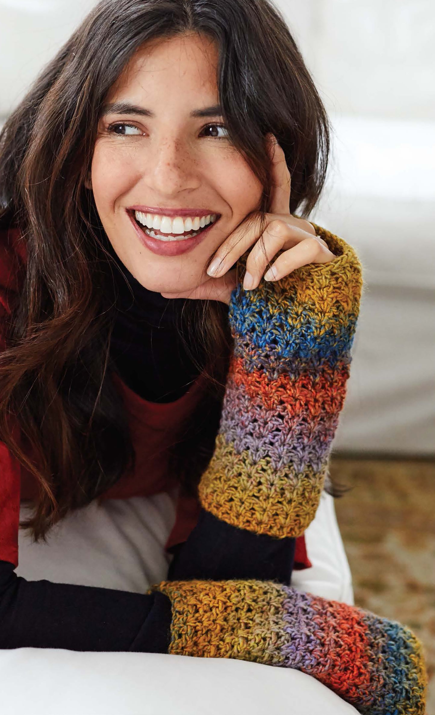 A smiling woman with long brown hair wears colorful, striped crochet fingerless gloves made from Cakery Crochet by Leisure Arts and sits on a couch, resting her chin on her hand.