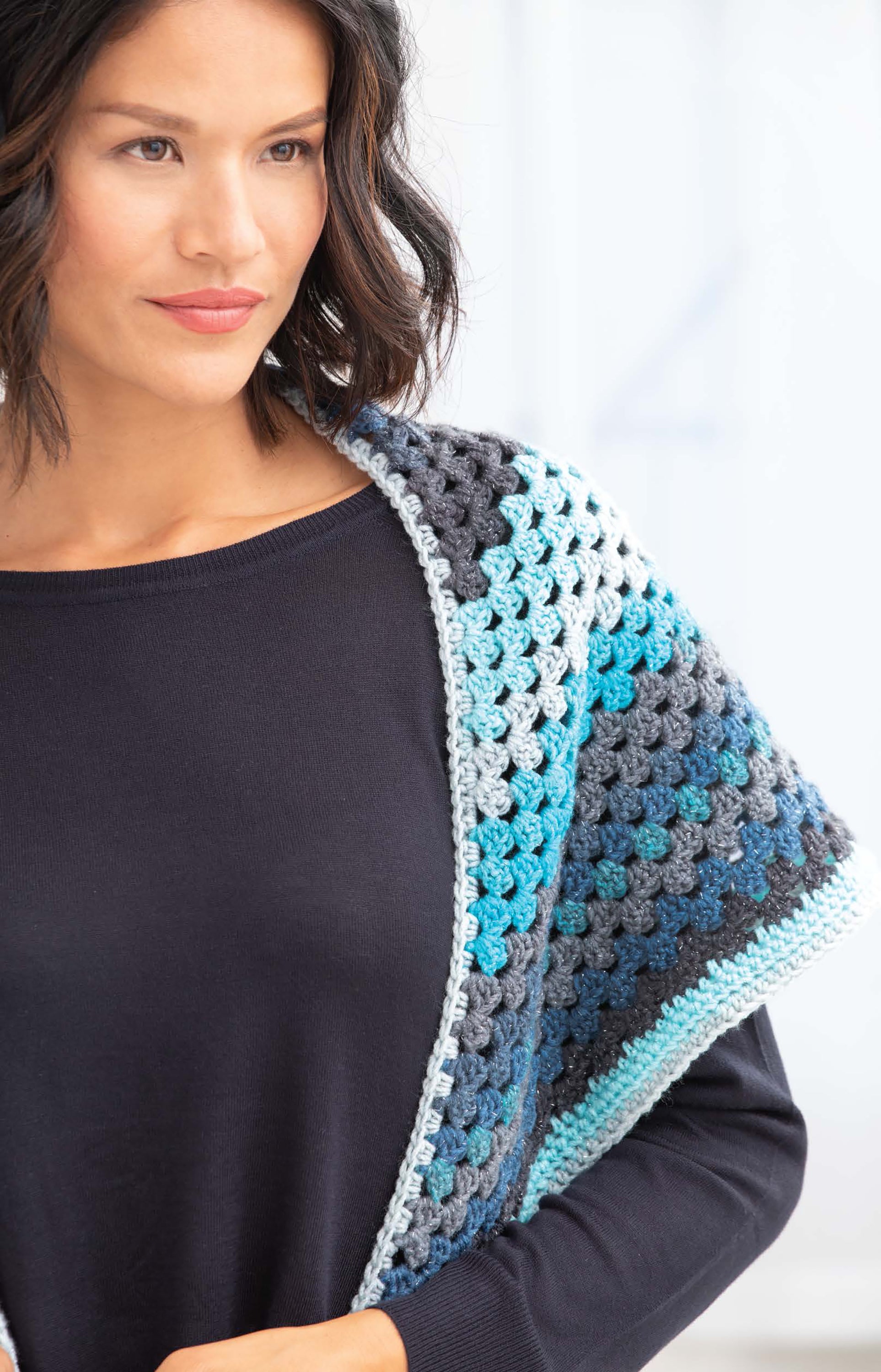 A woman with wavy dark hair wears a black top and a multicolored shawl made using Cakery Crochet by Leisure Arts in blue, grey, and white—an inspiring choice for beginners searching for easy crochet patterns.