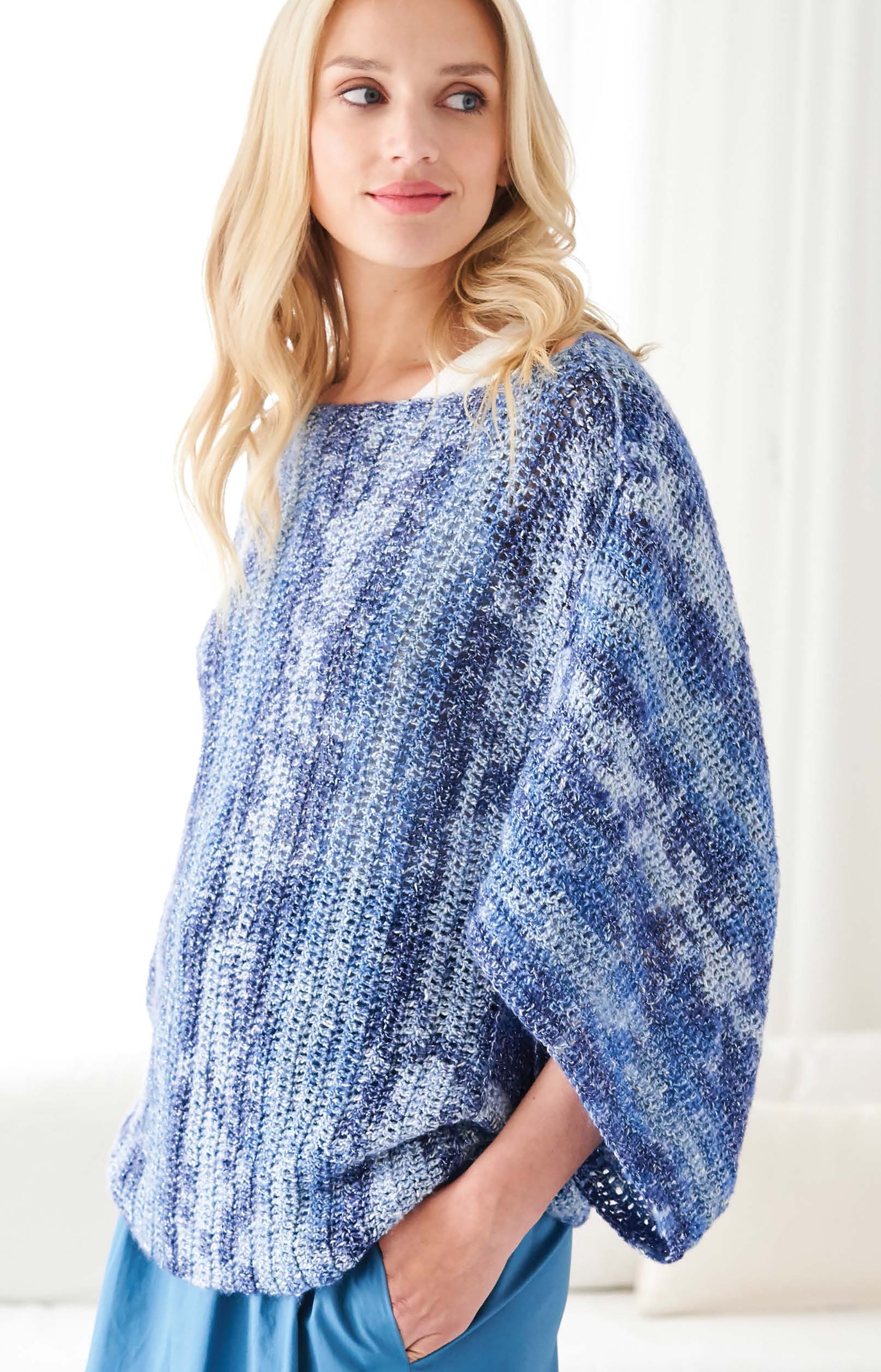 A woman with long blonde hair wears the Leisure Arts Cakery Crochet knit sweater in blue and white, paired with blue pants, as she stands in a bright room looking to the side with a gentle smile.
